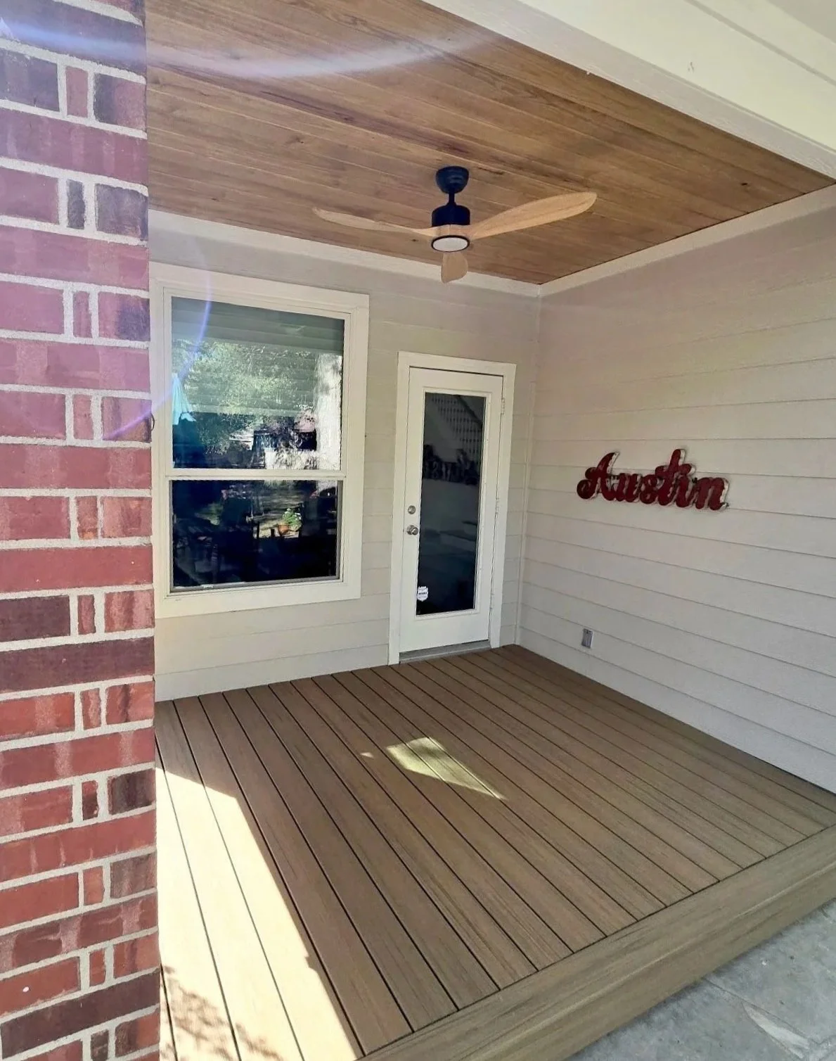 Covered porch with wooden flooring, a white door with glass, a window, a ceiling fan, and a decorative sign reading 'Austin' on the wall.
