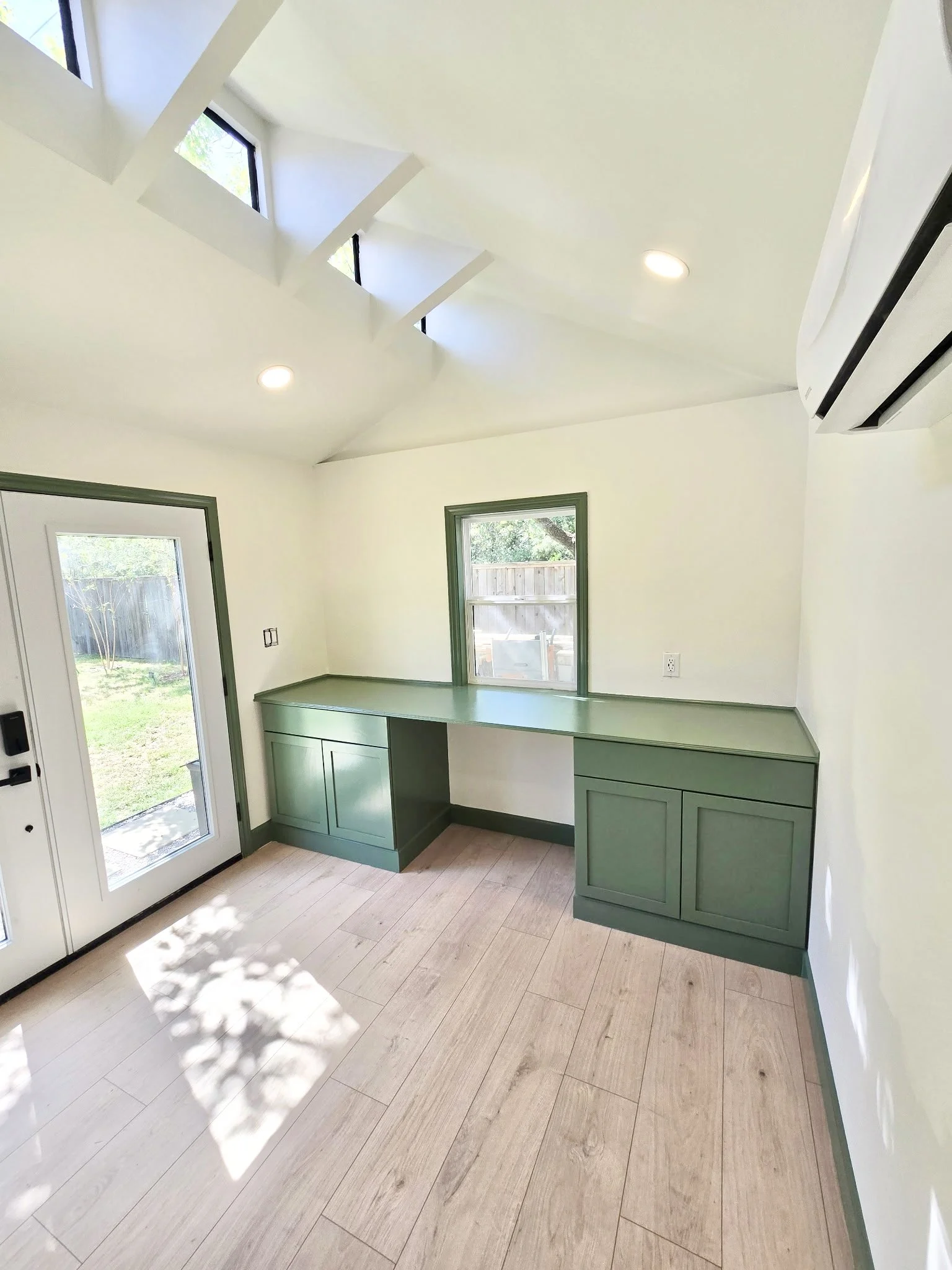 Interior view of a kitchen corner with light wood flooring, green cabinetry, white walls, and several windows letting in natural light.