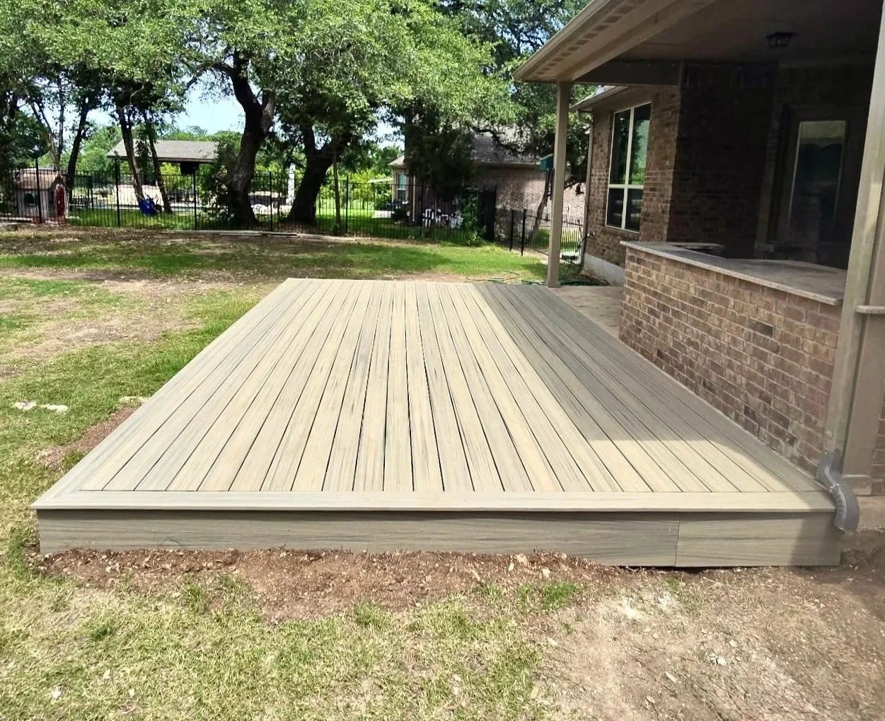 Backyard patio with newly installed wooden deck, adjacent to brick house with screened porch, surrounded by green grass, trees, and a fenced yard.