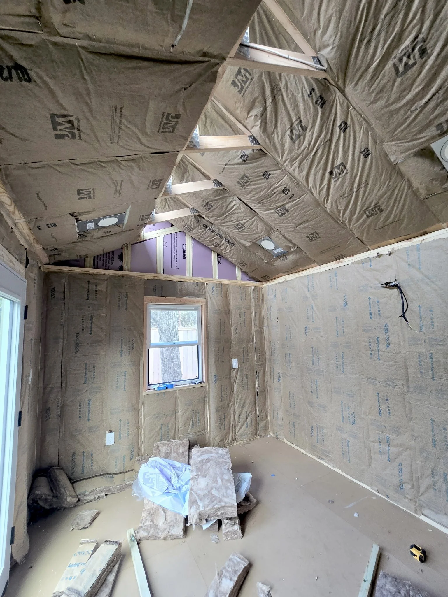 Room under construction with insulation material and drywall in progress, windows visible, and construction tools on the floor.