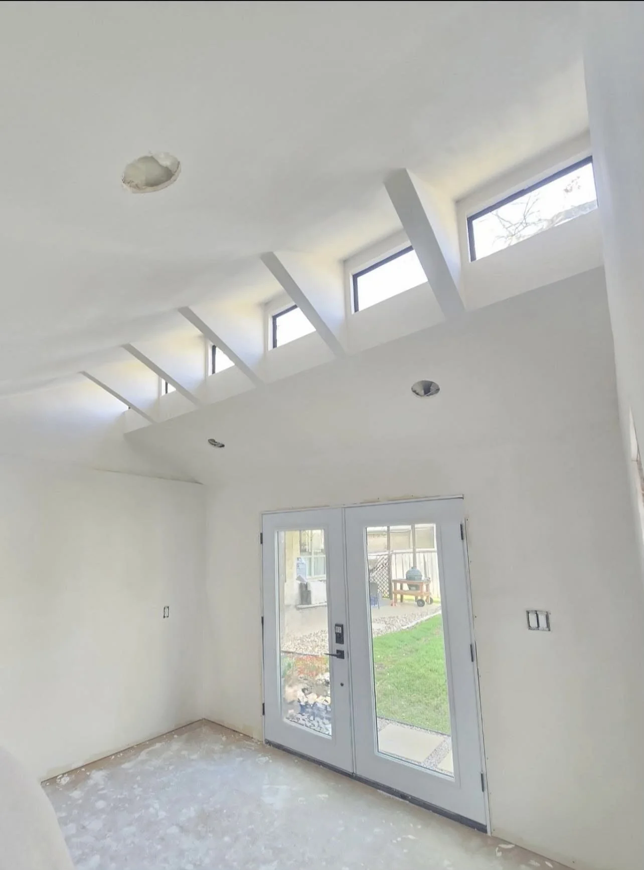 Interior of a room under construction with white walls and ceiling, large windows near the ceiling, and a glass door leading outside. The floor is unfinished, with some construction debris visible.