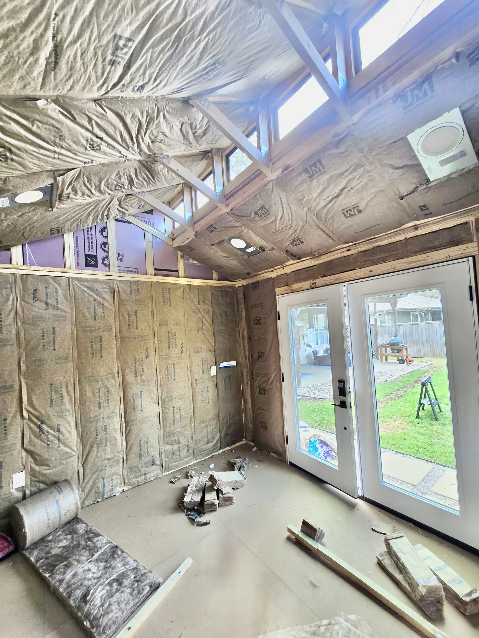 A room under construction with insulation and drywall installation in progress, with insulation materials on the walls and ceiling, a sliding glass door leading outside, and construction debris on the floor.