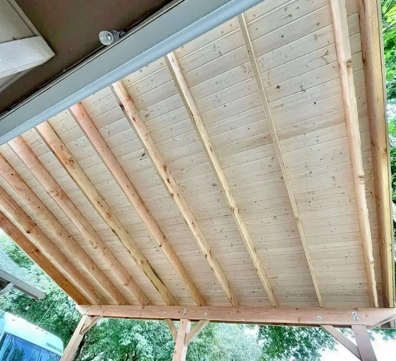 A wooden porch ceiling with exposed beams and wood paneling, outdoors with trees visible in the background.