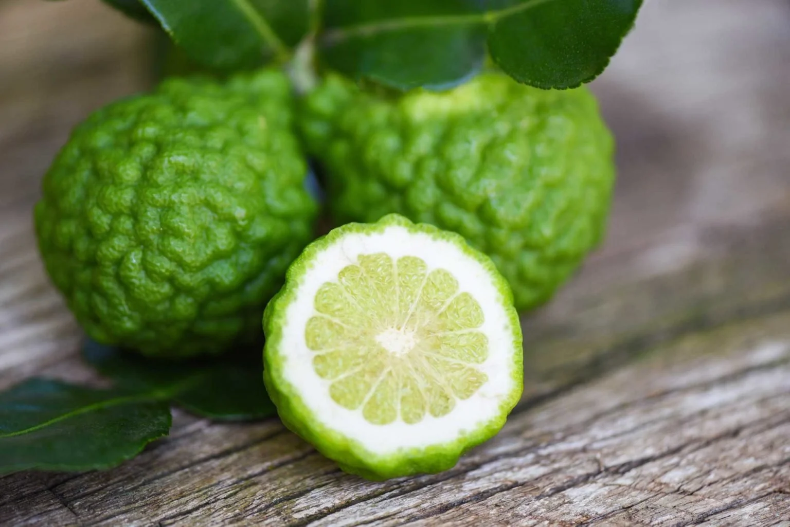 Bergamot fruit with one half cut, showcasing green, bumpy peel and interior, on a wooden surface with leaves.