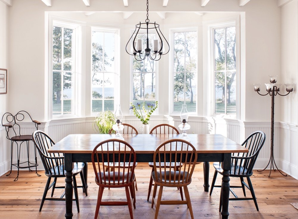 Bright dining room with a large wooden table, six mismatched chairs, a chandelier, a candelabra, and large windows with a view of a green outdoor landscape.