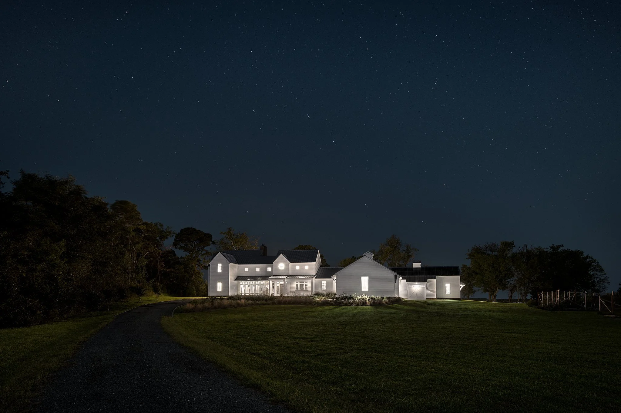 A large white house with lights on inside at night, set in a green yard with a dark walking path, trees on the left side, and a starry sky overhead.