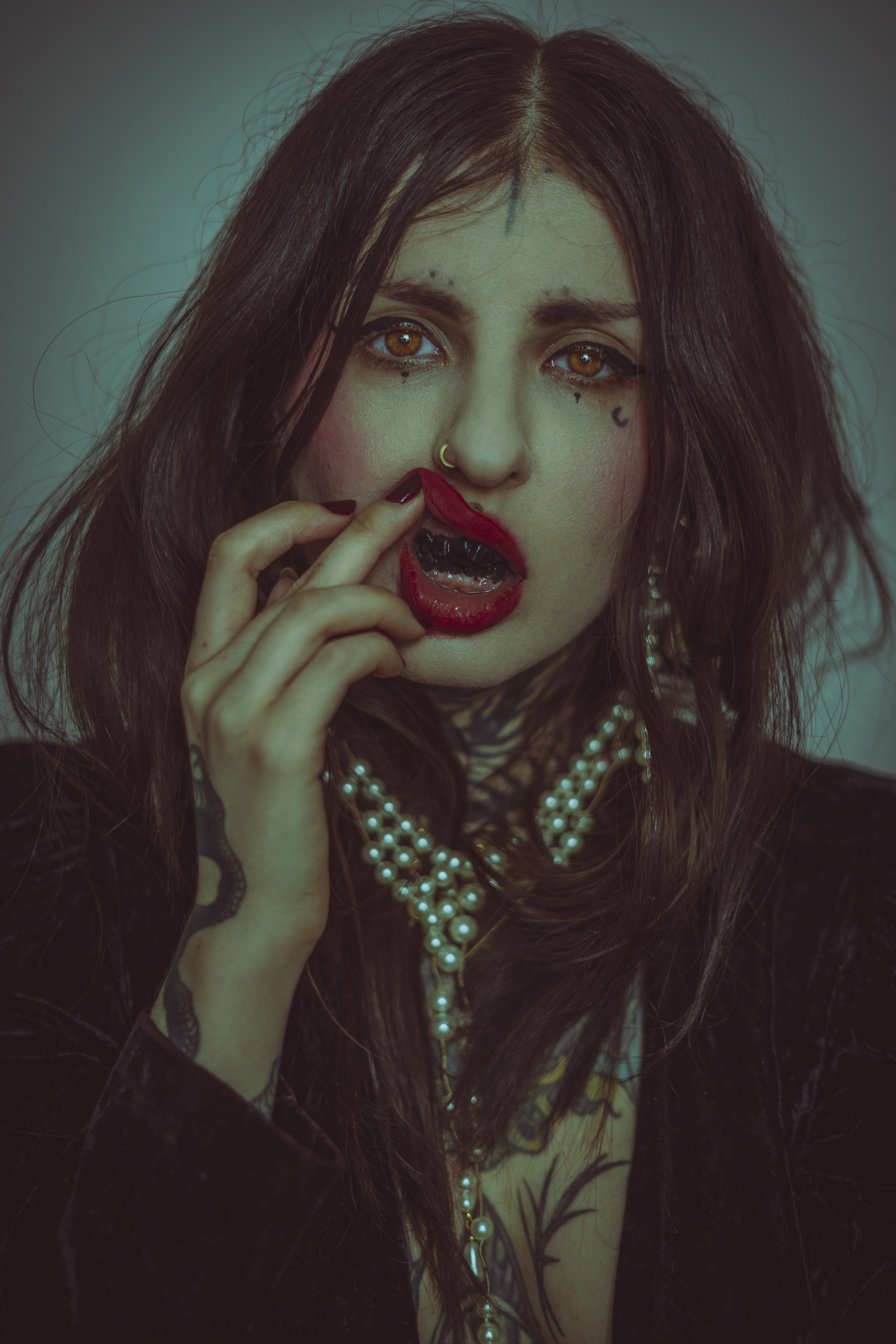 A woman with long brown hair and blue eyes is touching her lips with her fingers, wearing red lipstick and a pearl necklace, with tattoos visible on her hand and neck.