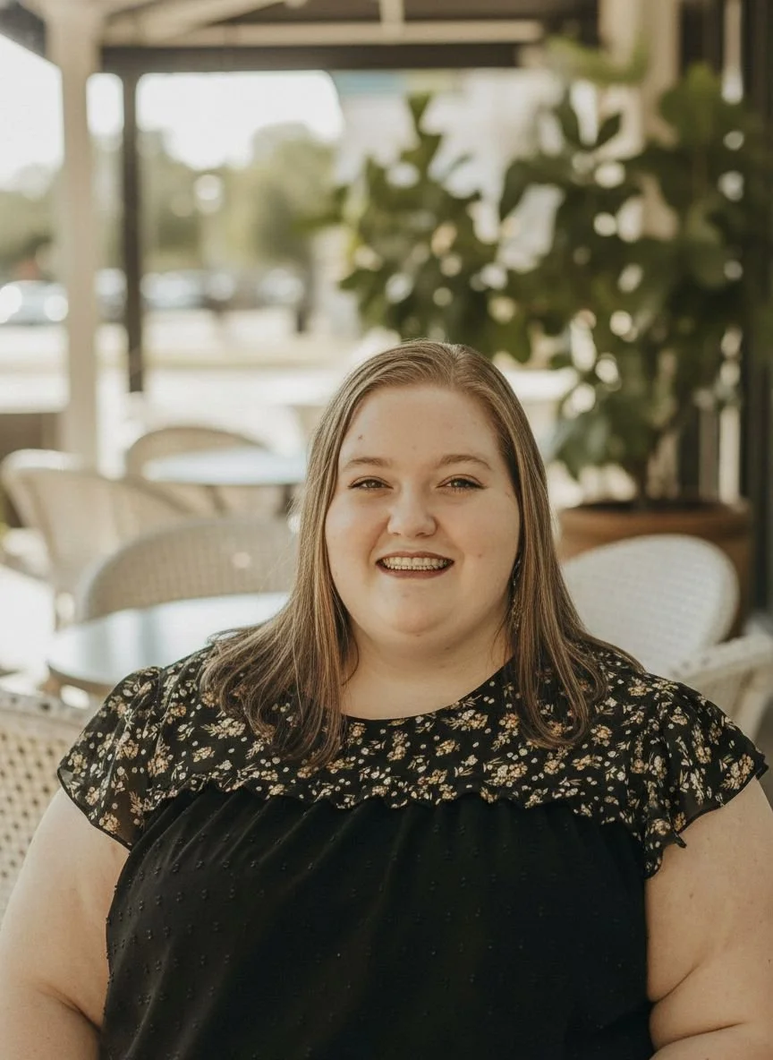 Sam with shoulder-length brown hair, smiling, wearing a black top with a floral patterned yoke and ruffled sleeves, sitting in a casual cafe or restaurant setting with wicker chairs and large windows, natural light, and greenery outside.