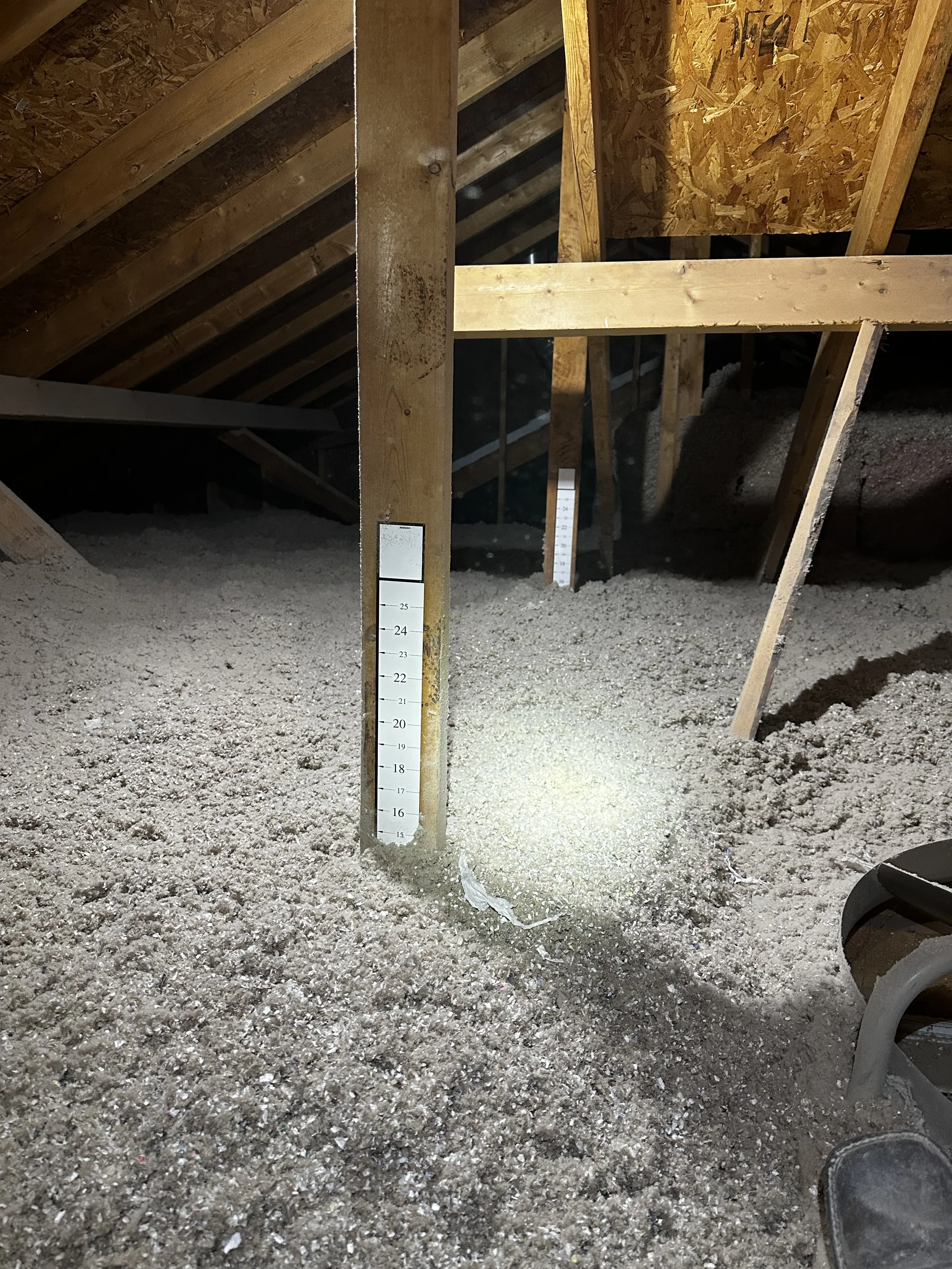 Interior of an attic with exposed wooden beams and a dust-covered floor, featuring a vertical measuring stick attached to a wooden post for height reference.