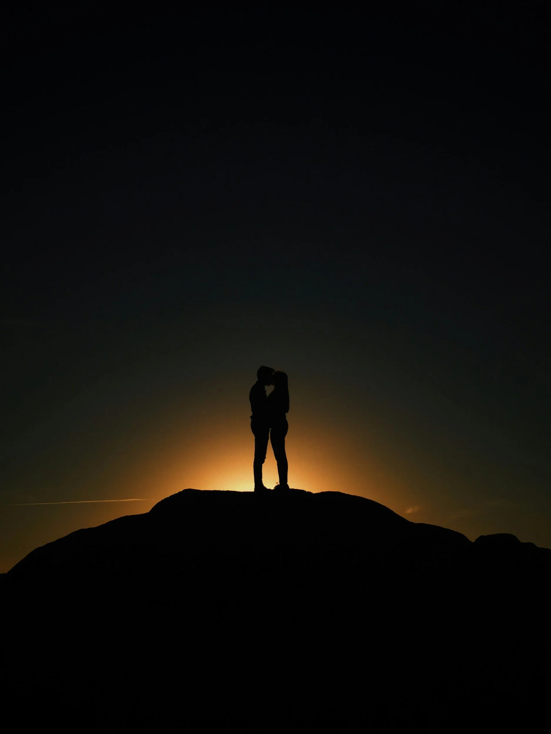 Silhouette of a couple standing on a hilltop kissing, with a sunset or sunrise in the background creating a glowing halo around them.