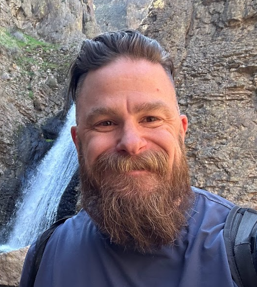 A man with a beard and mustache taking a selfie outdoors near a waterfall and rocky terrain.  sex therapy in Utah or Idaho.