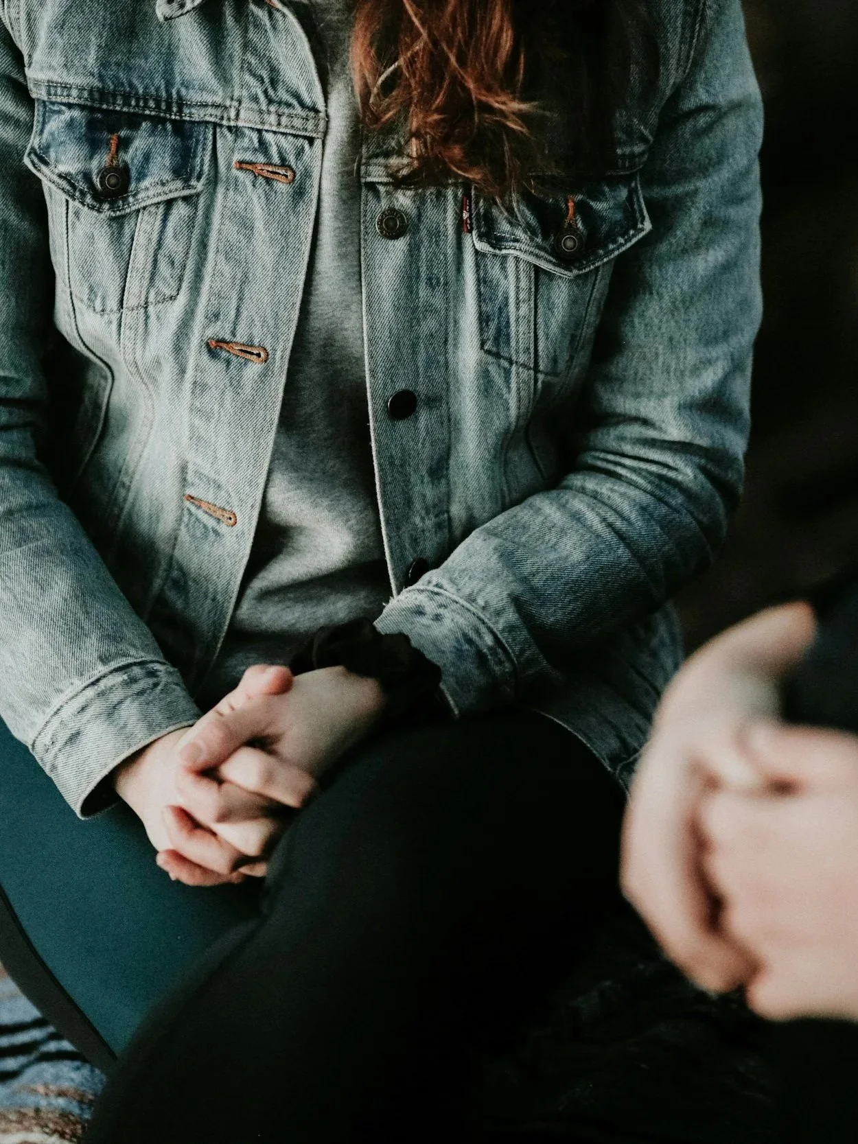 Person wearing a blue denim jacket and gray sweatshirt sitting with hands clasped, holding a smartphone.