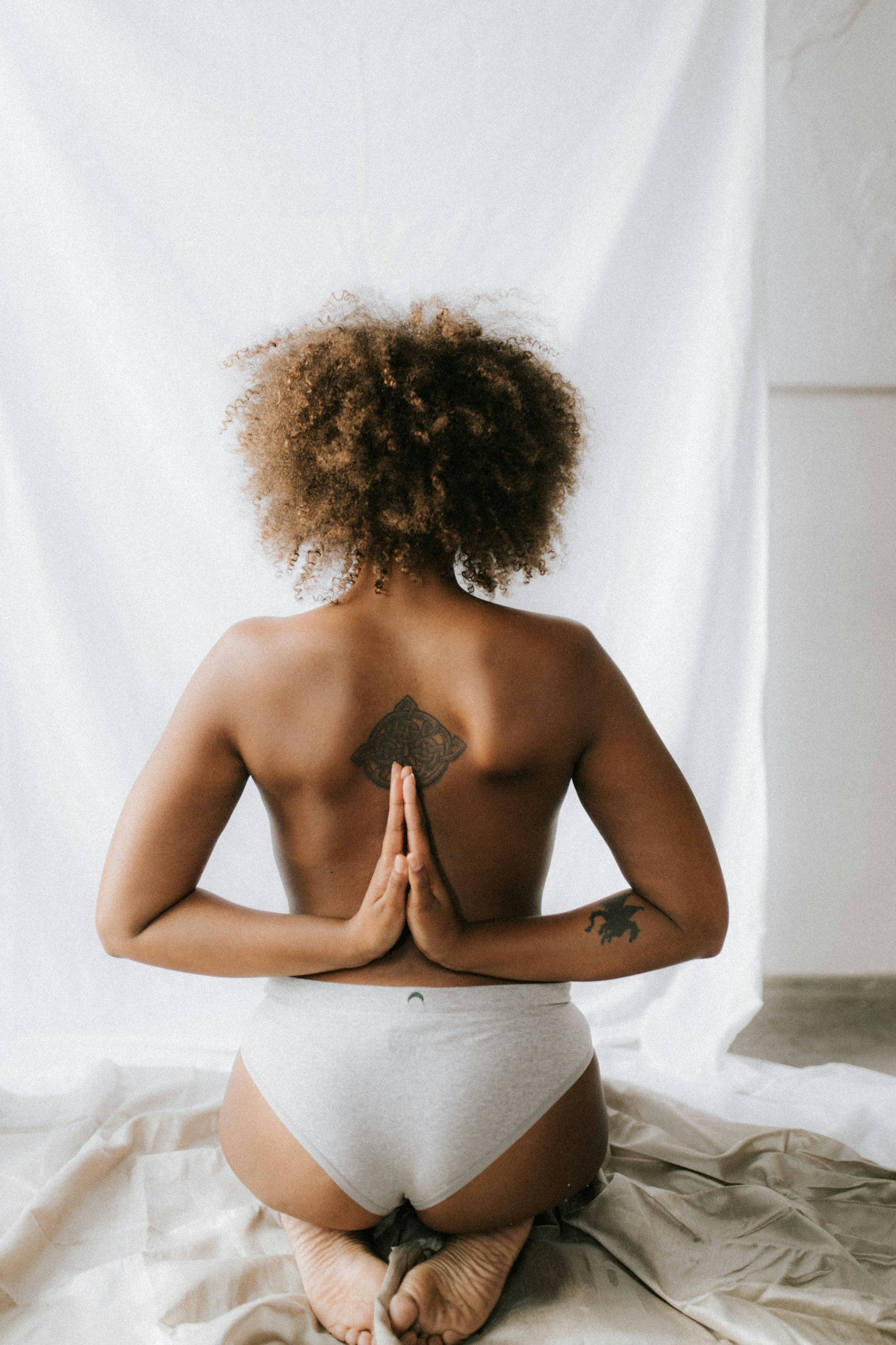 A woman with curly hair kneeling on the floor with her back to the camera, placing her hands together behind her back in a prayer pose, wearing gray bottoms, with a tattoo on her upper back and another on her left arm.