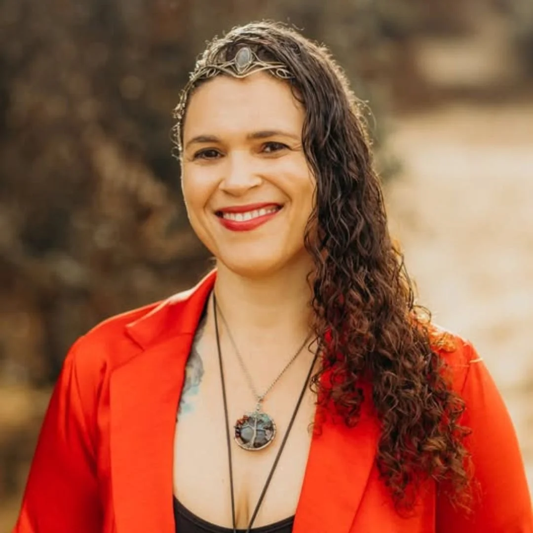 A woman with long, curly dark hair wearing a red blazer, red lipstick, and a necklace with a circular pendant, smiling outdoors with blurred background.