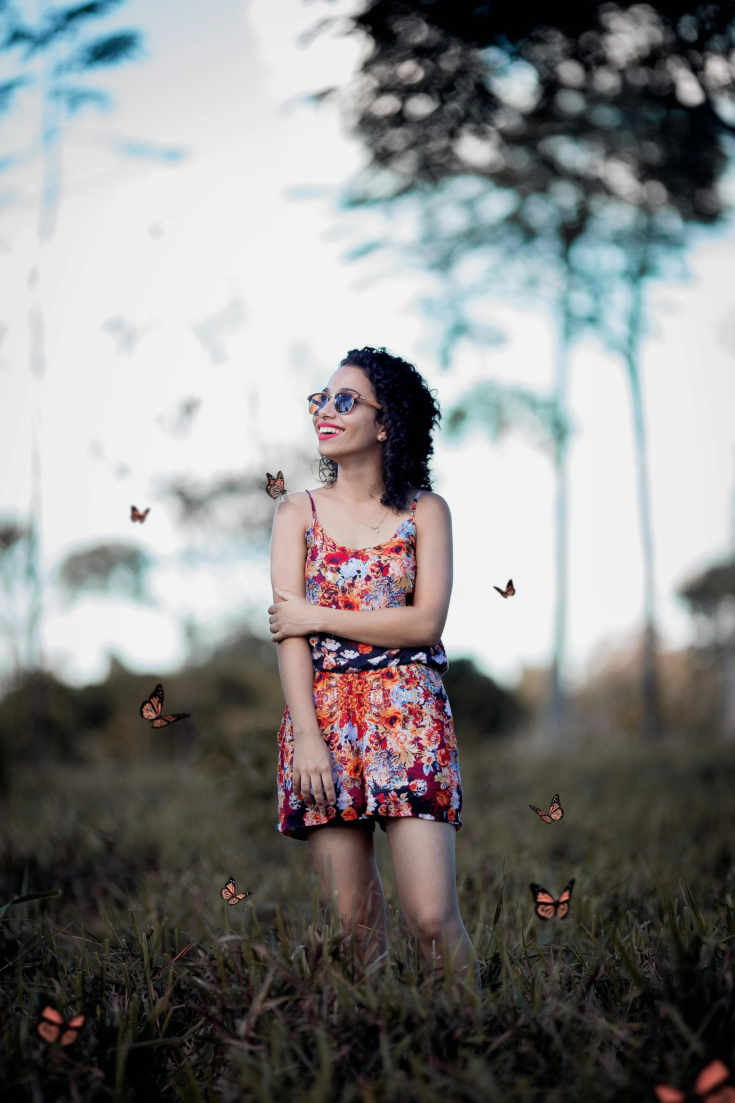 A woman with curly hair and sunglasses wearing a floral dress standing in a grassy outdoor area with butterflies around her, trees in the background, smiling and looking upward.