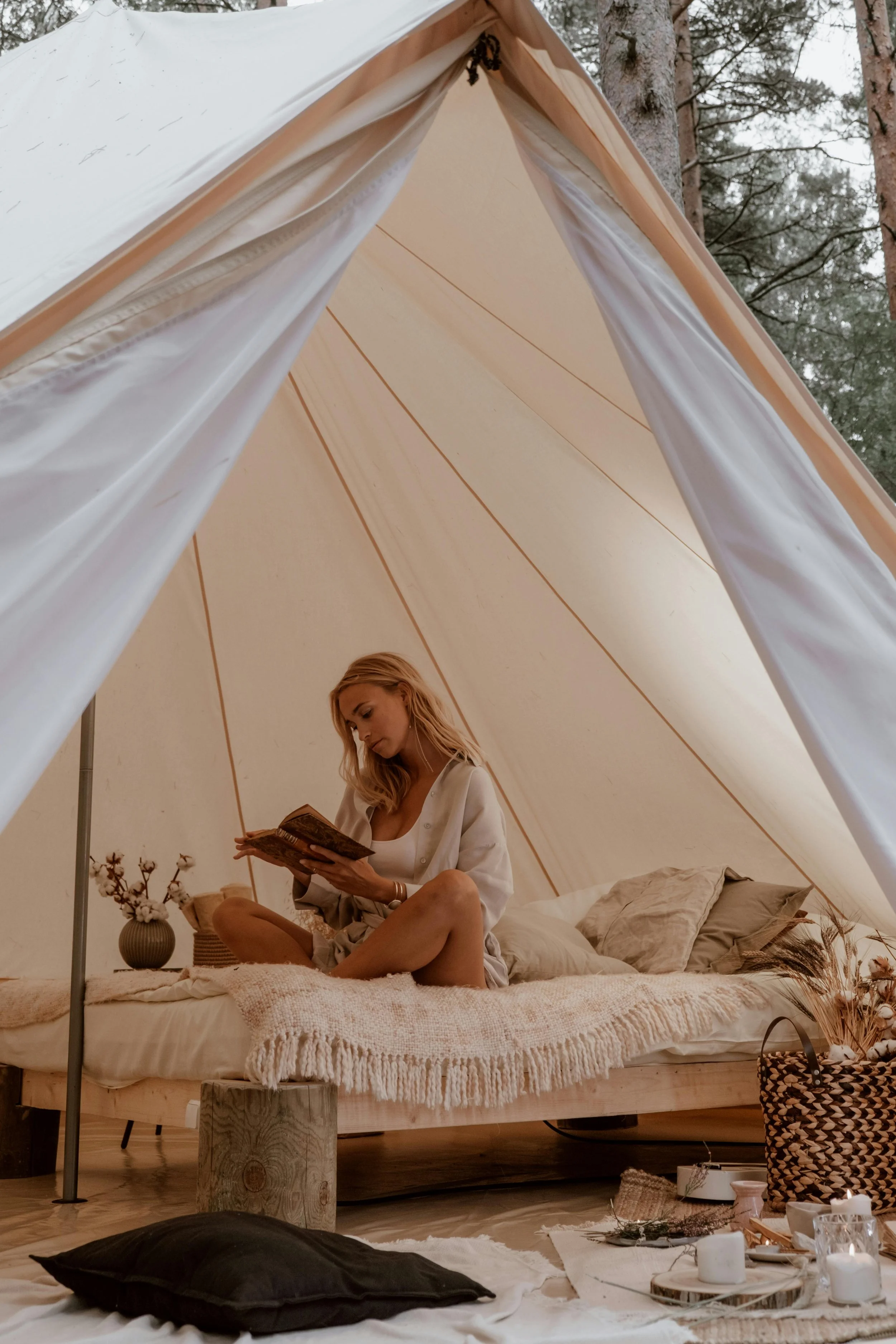 A woman sitting inside a cozy beige camping tent, reading a book, with pillows, a small vase with flowers, and various candles and decorations around her.