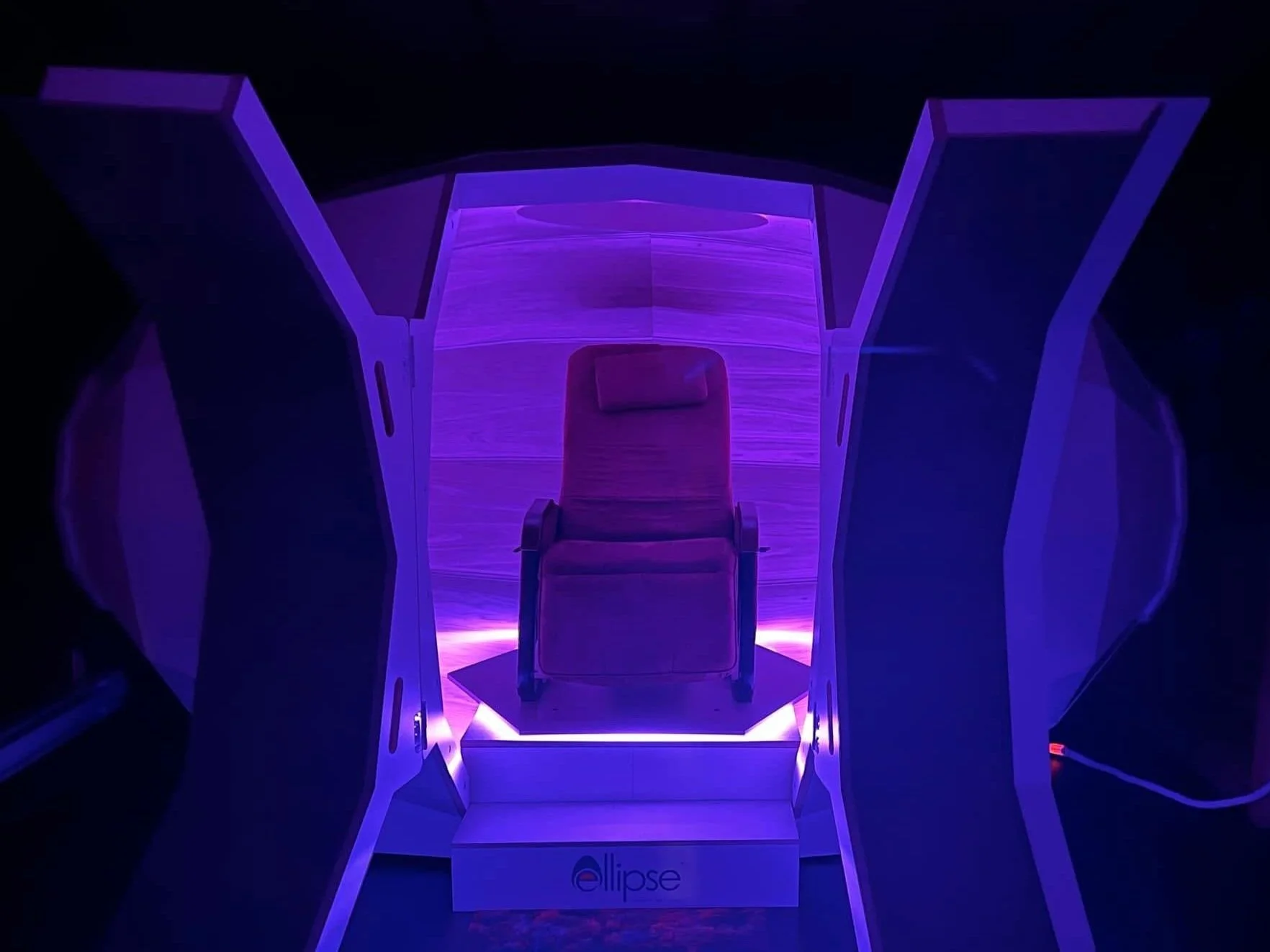 A chair inside a high-tech device with purple lighting, surrounded by large panels that are open, with a wooden background and the logo 'ellips' at the base.