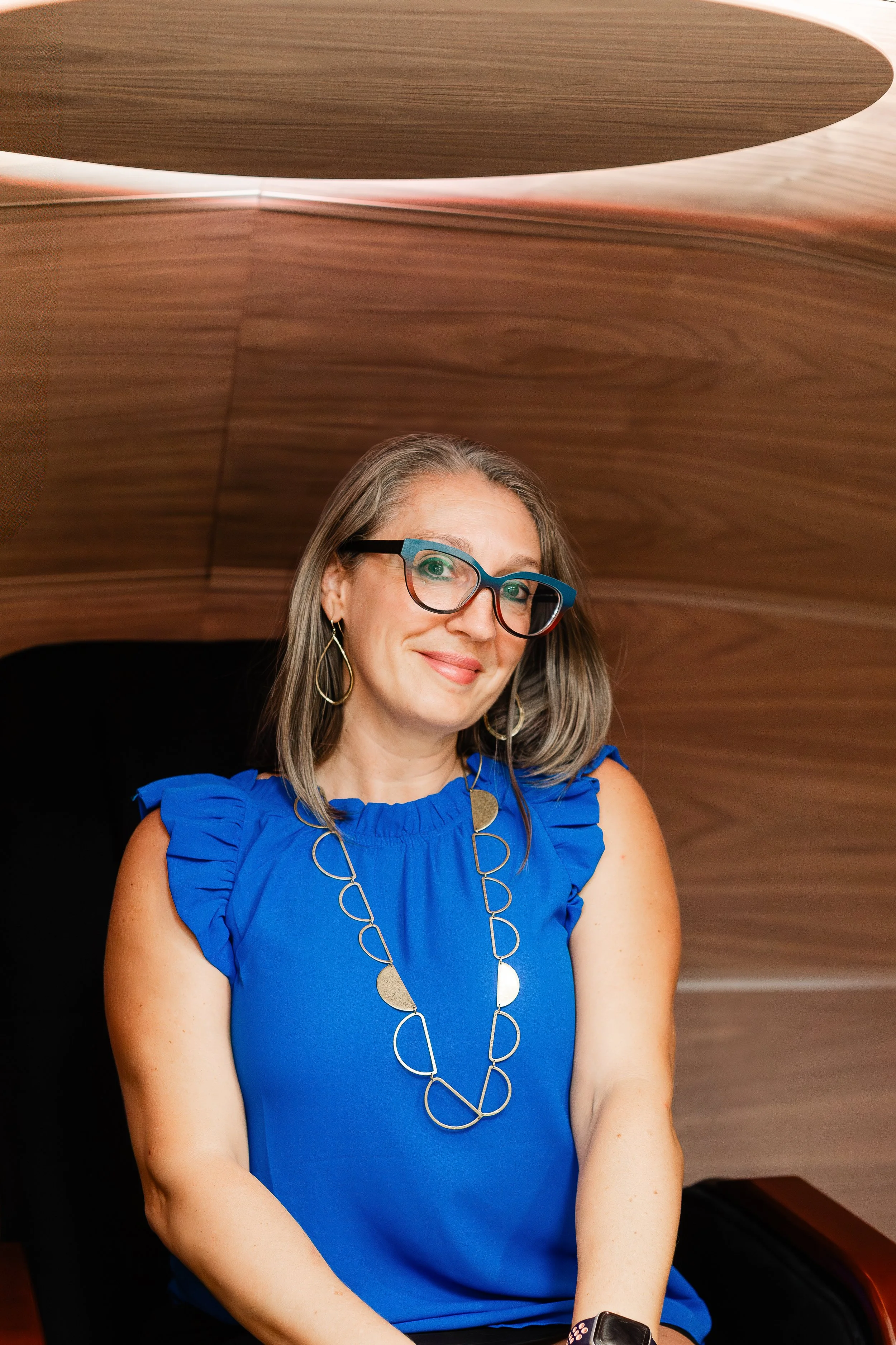 A woman wearing glasses and a blue ruffled sleeveless blouse sitting in front of a wooden background.