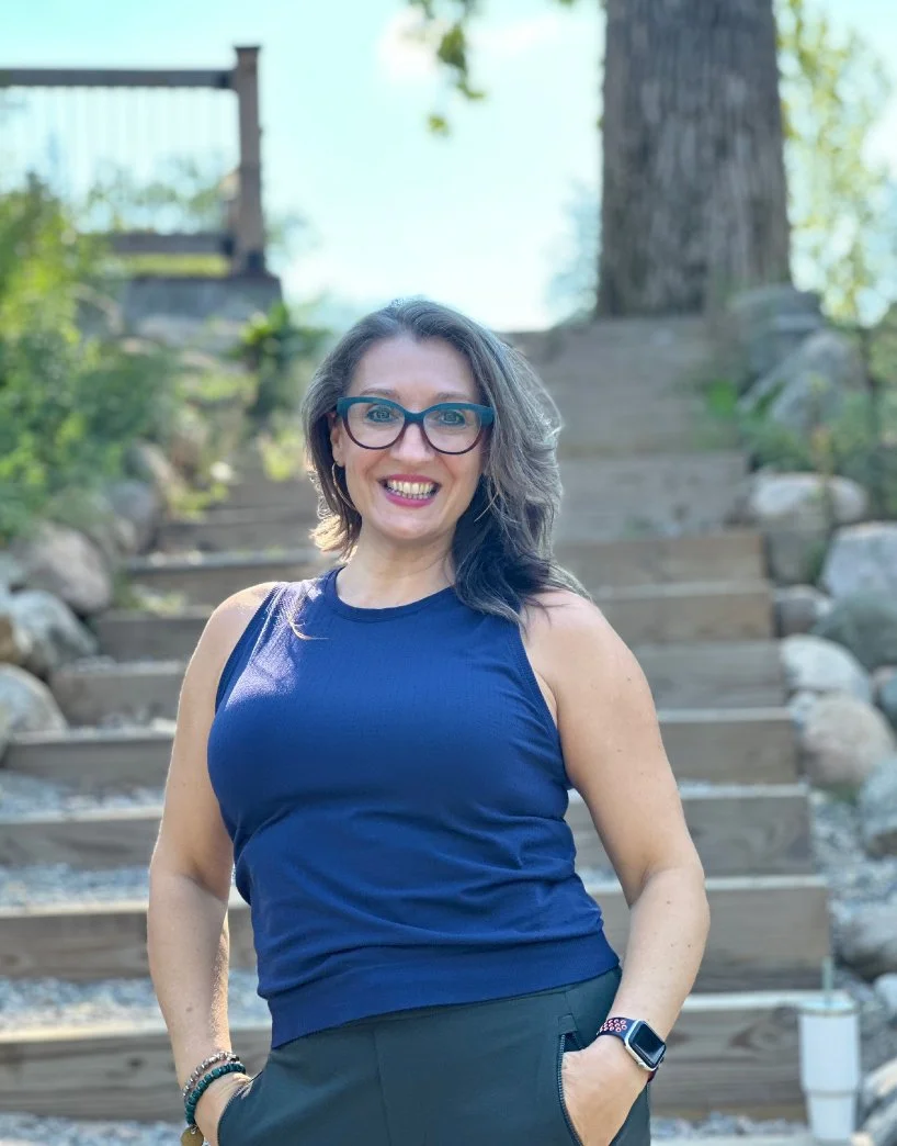 A woman with glasses and shoulder-length gray hair smiling outdoors, standing in front of a wooden staircase and green trees.