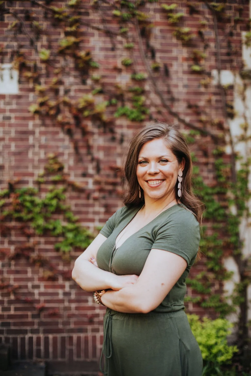A woman with brown hair wearing an olive green dress with short sleeves, standing outdoors in front of a brick wall with green vines, smiling and crossing her arms.