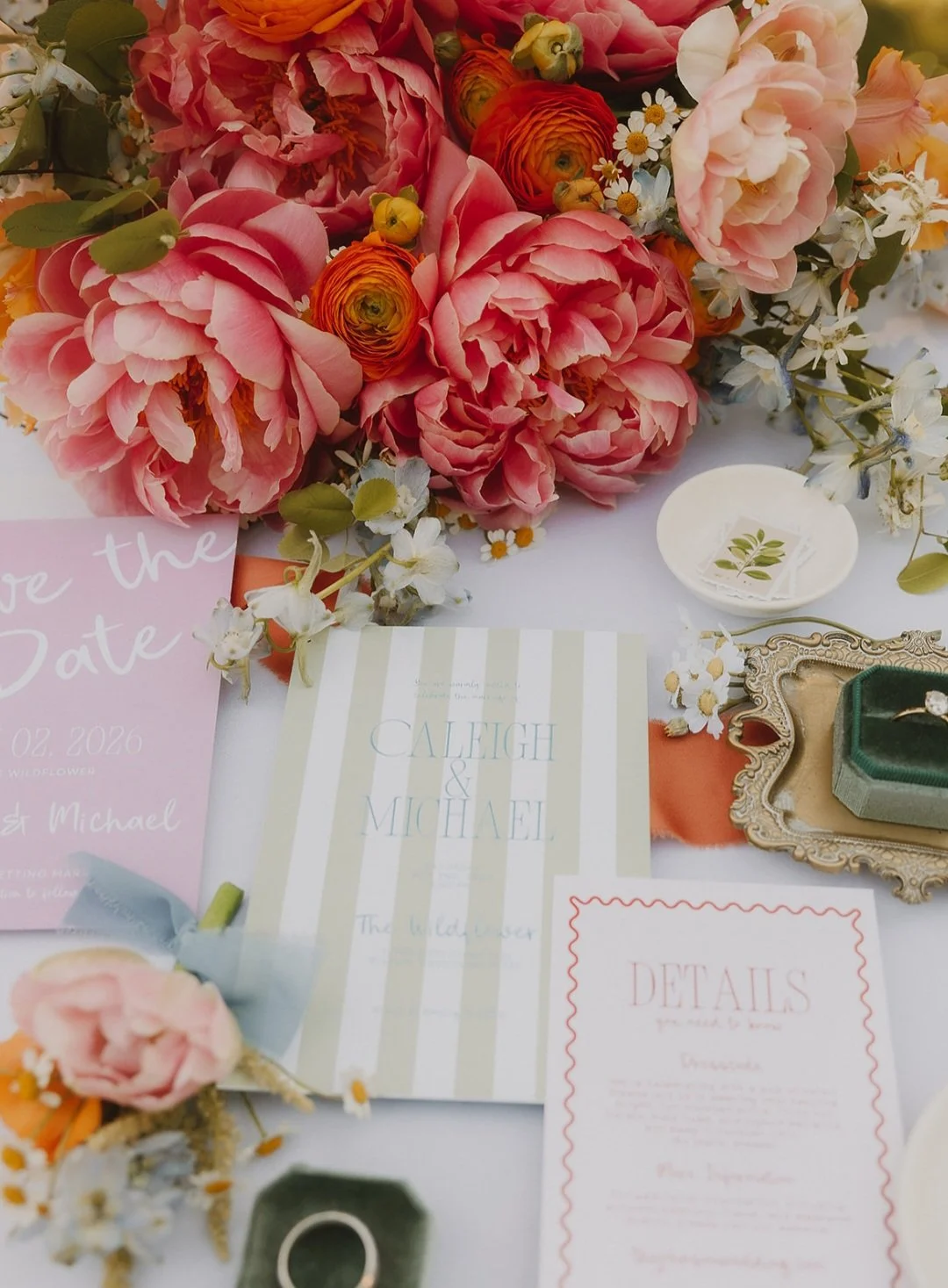 Close-up of a floral arrangement with pink and orange peonies, ranunculus, daisies, and other small white flowers on a wedding table with invitations and decorative items.