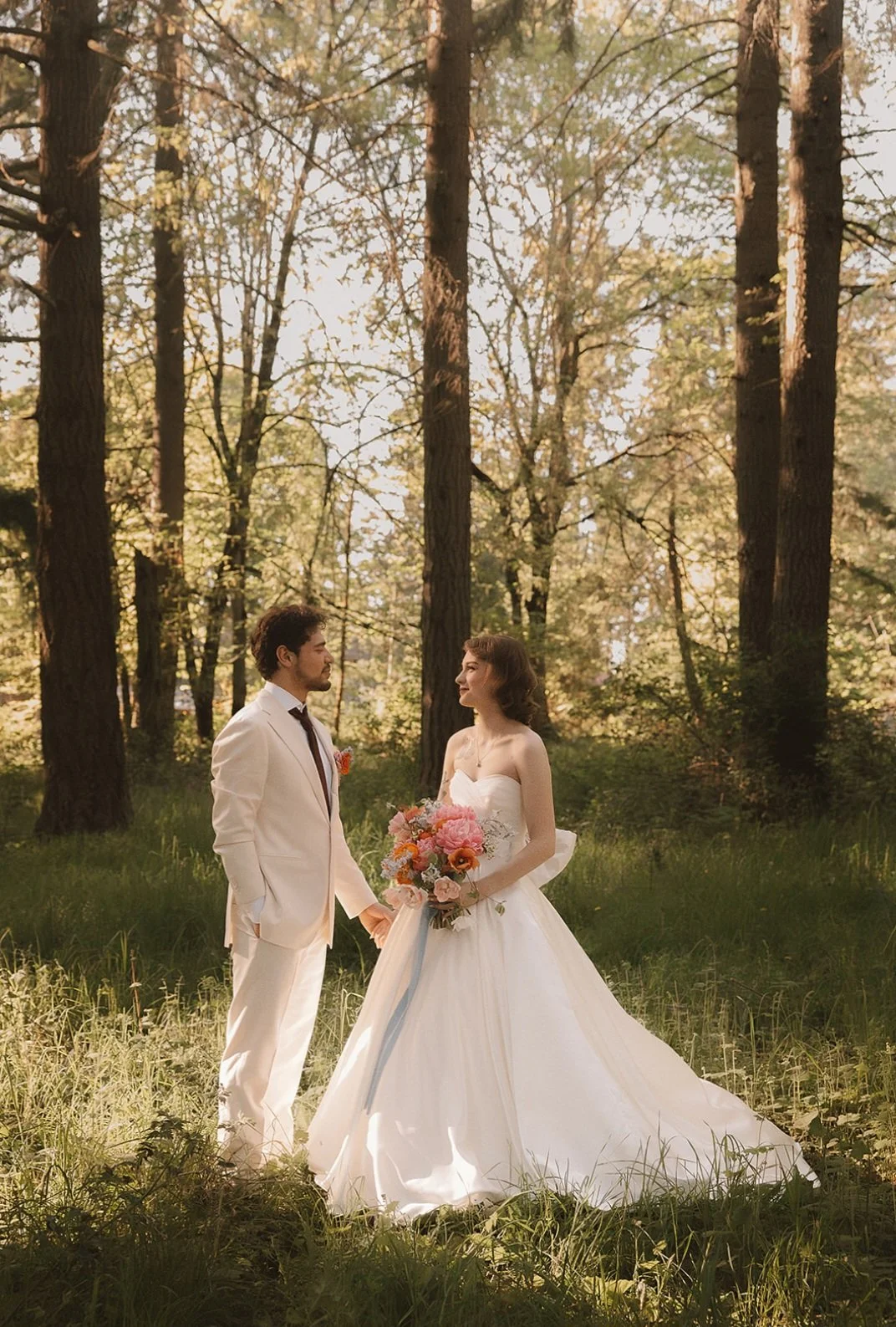 A newlywed couple standing hand in hand in a forest clearing. The groom is in a white tuxedo with a purple tie, and the bride is in a strapless white wedding gown holding a bouquet of pink and orange flowers. They are looking at each other amidst tal