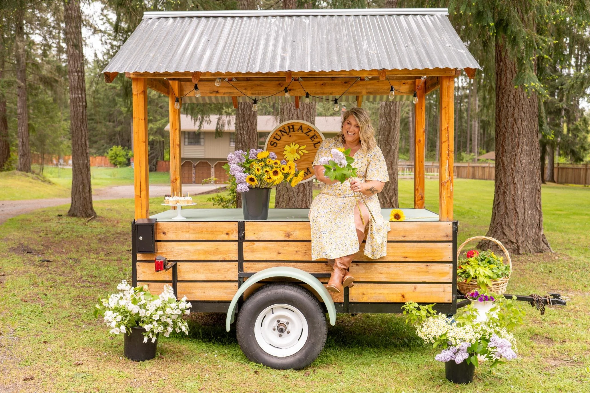 A woman with blonde hair in a floral dress sitting on a wooden cart decorated with colorful flowers, located outdoors in a wooded area.