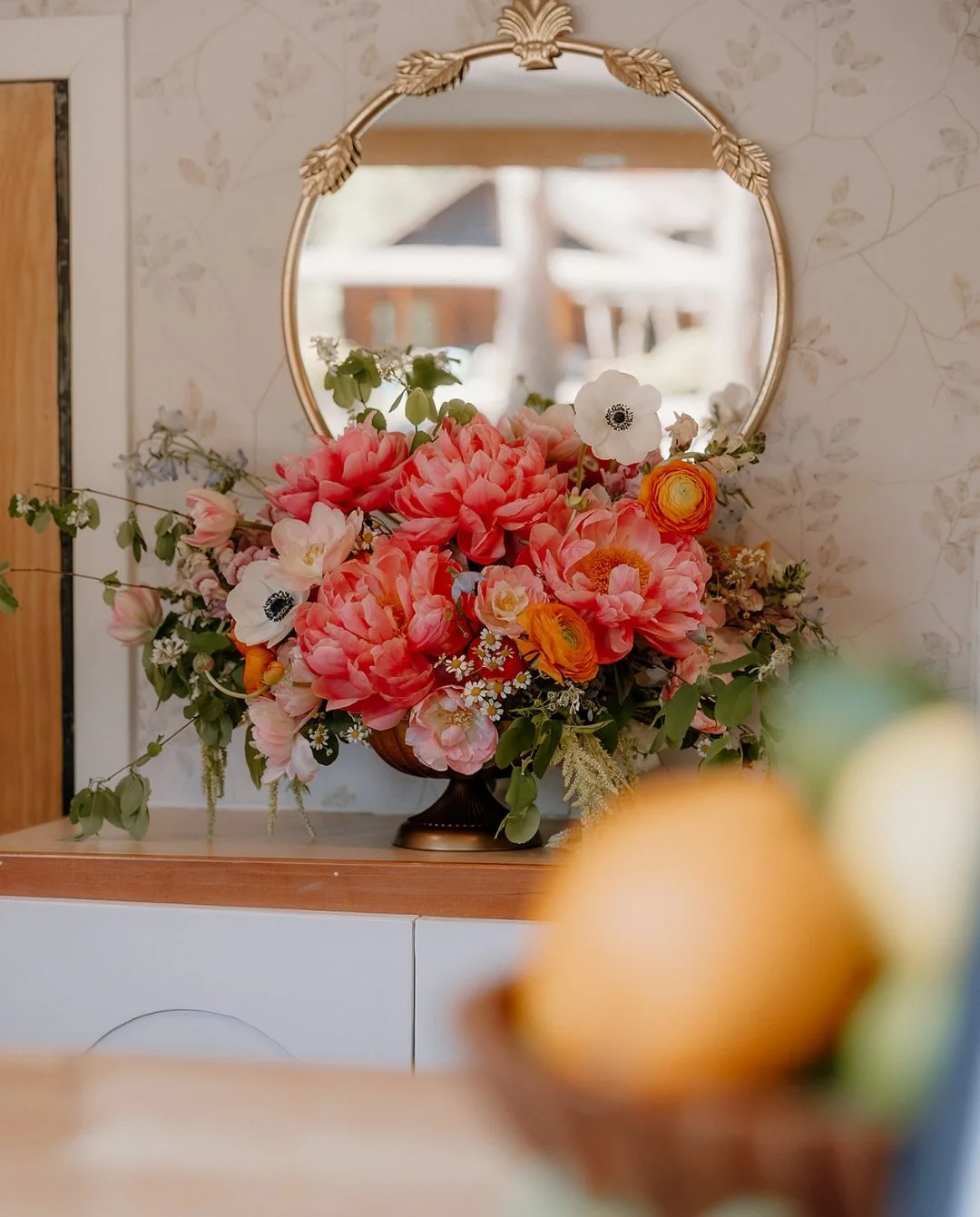 A floral arrangement with pink, white, and orange flowers in a dark brown vase, reflected in an ornate gold-framed mirror on a wooden surface against a wallpapered wall.