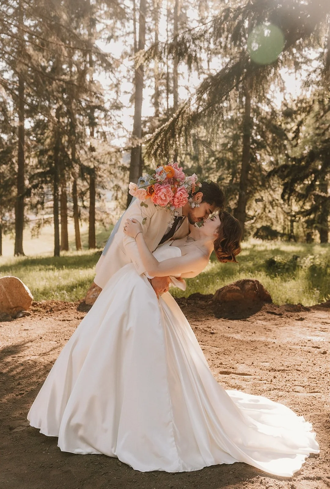 A bride and groom in wedding attire sharing a romantic moment outdoors surrounded by trees, with the groom holding the bride in a dip, and the bride holding a bouquet of pink and orange flowers.