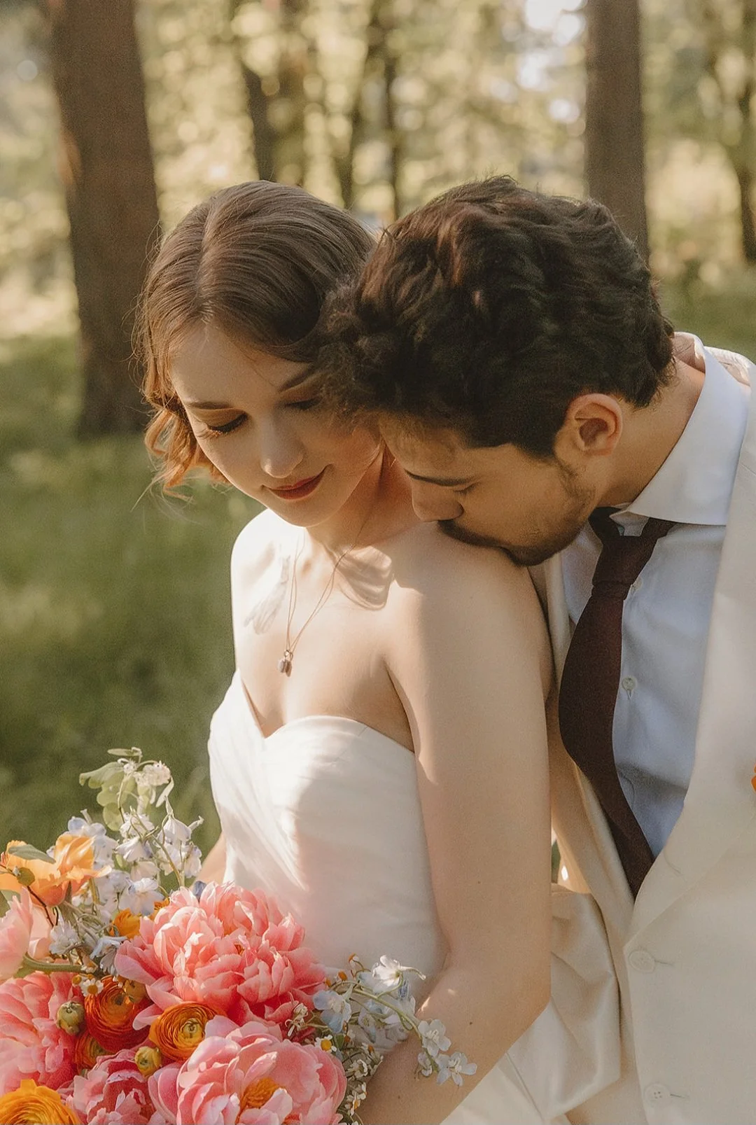 A bride and groom sharing an intimate moment outdoors with trees in the background, the bride holding a colorful bouquet of flowers.