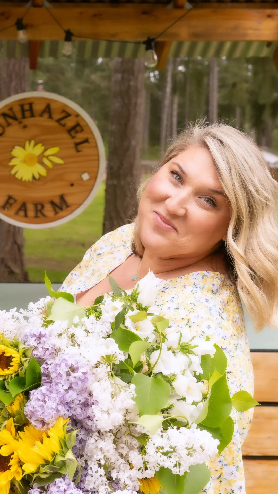 A woman with blonde hair holding a large bouquet of white, purple, and yellow flowers, standing outdoors beside a wooden sign that reads 'HONE AZEL FARM' with a yellow flower illustration.