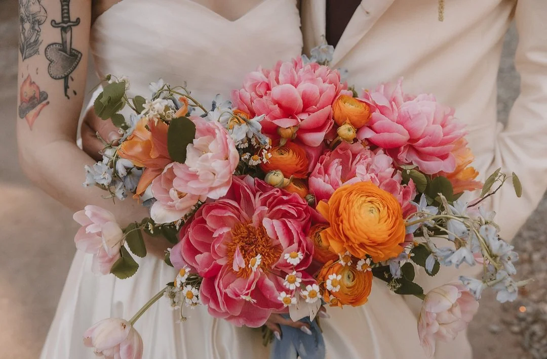 Bride holding a bouquet of pink, orange, peach, and light blue flowers including peonies, ranunculus, and daisies.