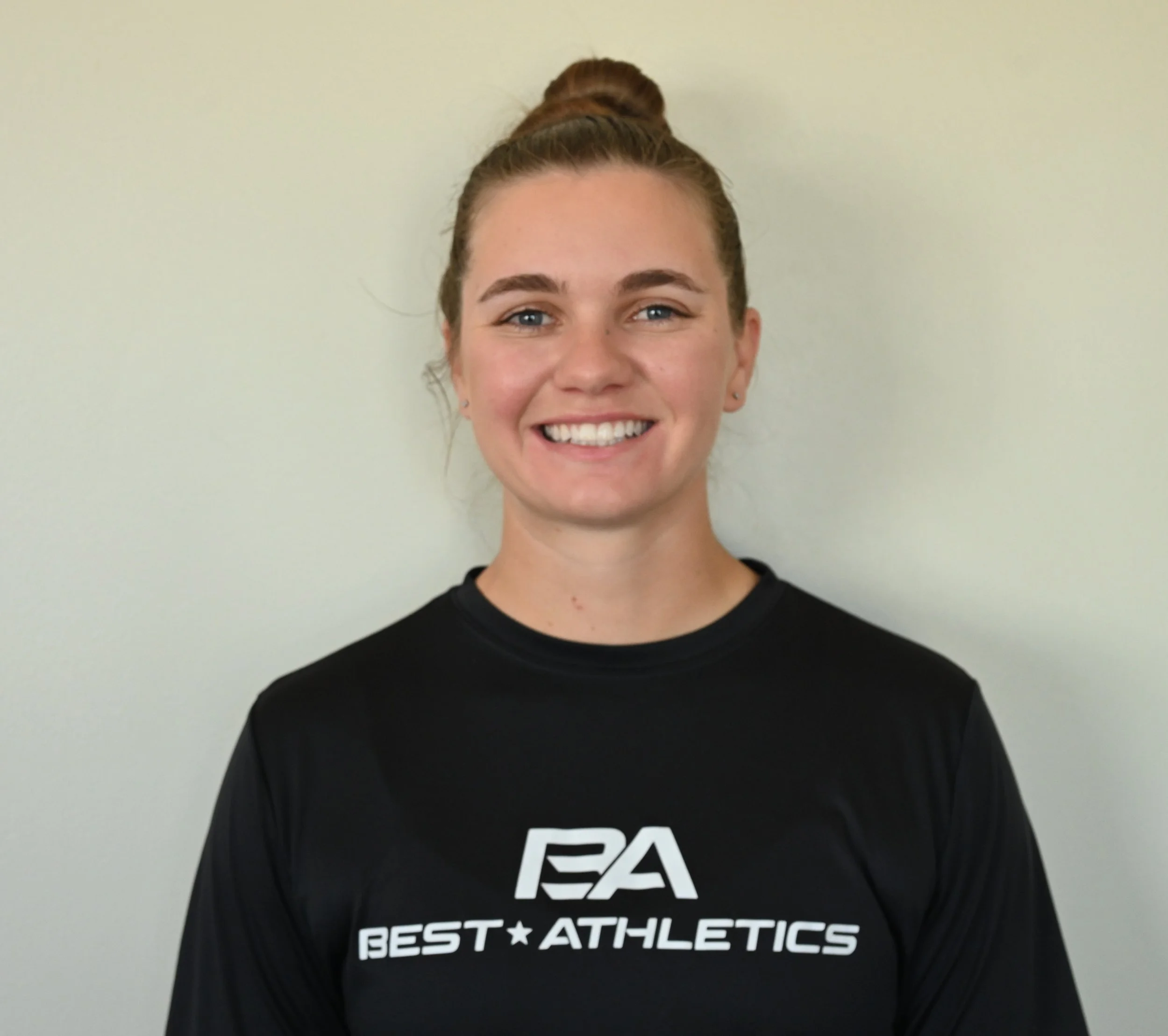 Close-up of a young woman with light skin and both brown and red hair styled in a high bun, smiling in front of a plain wall. She is wearing a black T-shirt with white text that reads, 'BEST ATHLETICS.'