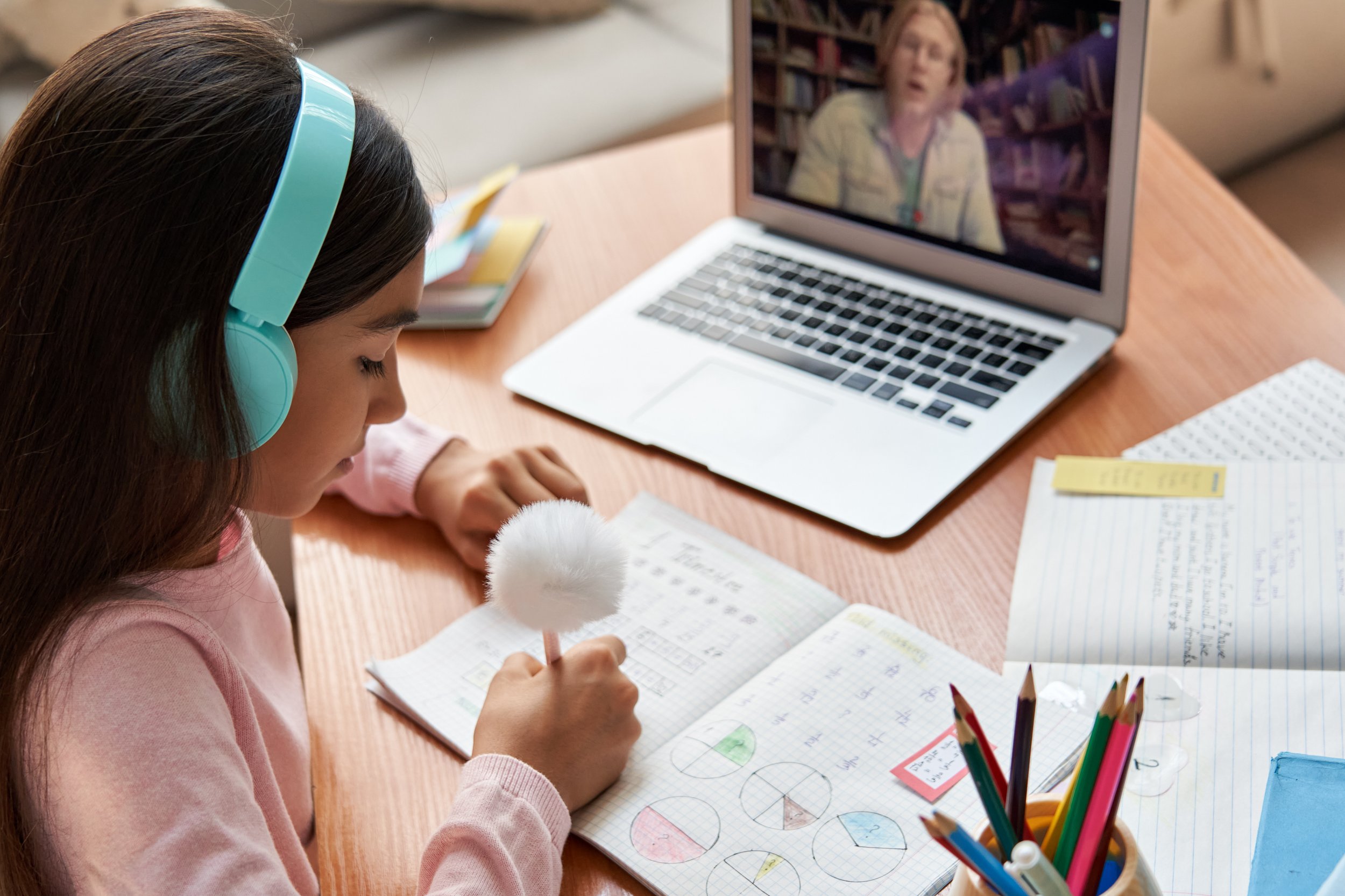 A girl wearing teal headphones studies with a notebook, microphone, and colored pencils at a desk while watching an online lesson on a laptop.