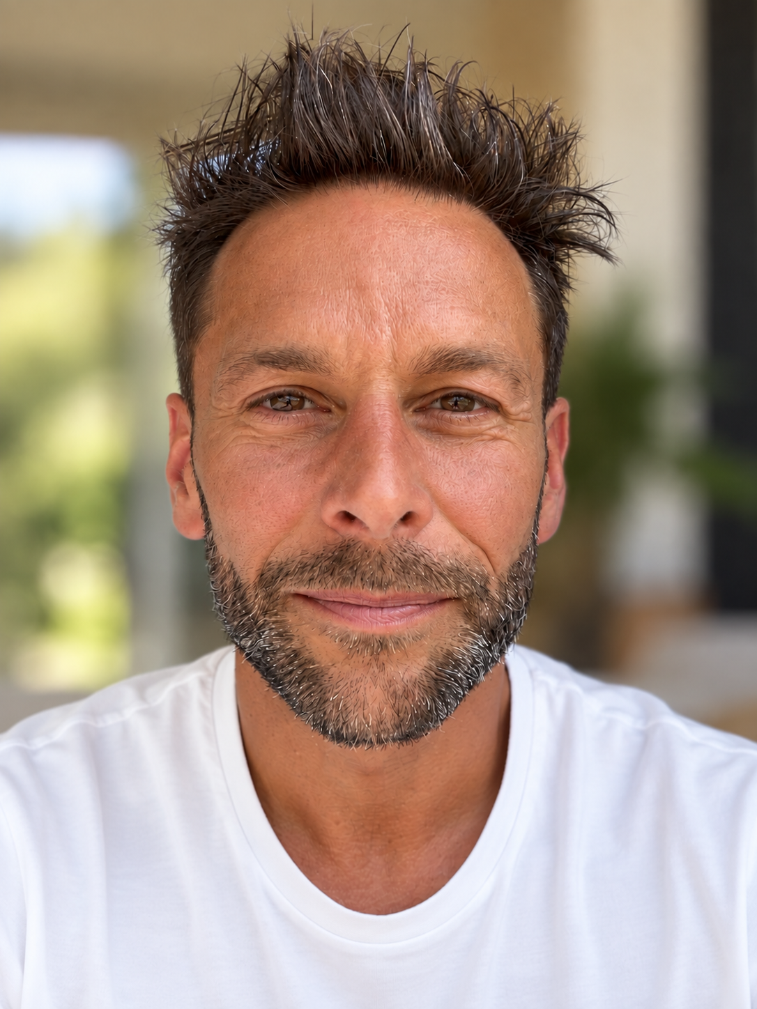 Un homme souriant avec une barbe brune, portant un t-shirt blanc, dans un environnement lumineux, vraisemblablement en extérieur ou dans une pièce bien éclairée.