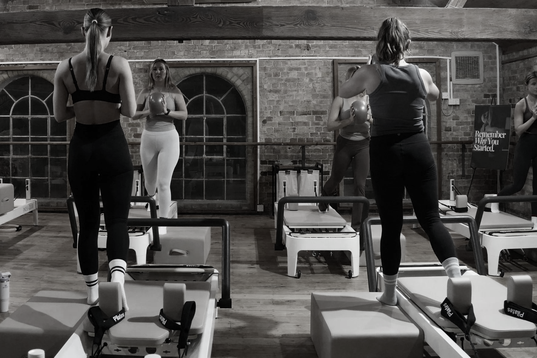 Group of women in a Pilates class, standing on Pilates reformer machines, holding kettlebells, in a gym with brick walls and large windows.