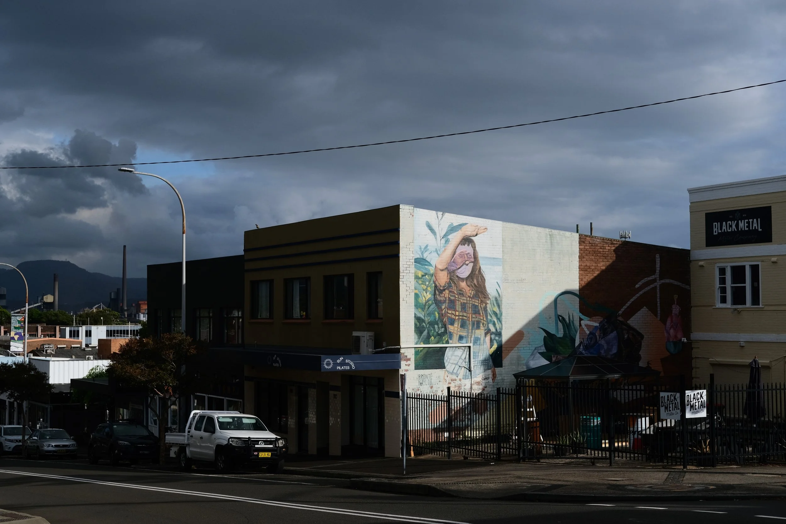 A mural on the side of a building depicts a woman with long hair wearing a plaid shirt and face mask, with a rural background, against a backdrop of a cloudy sky.