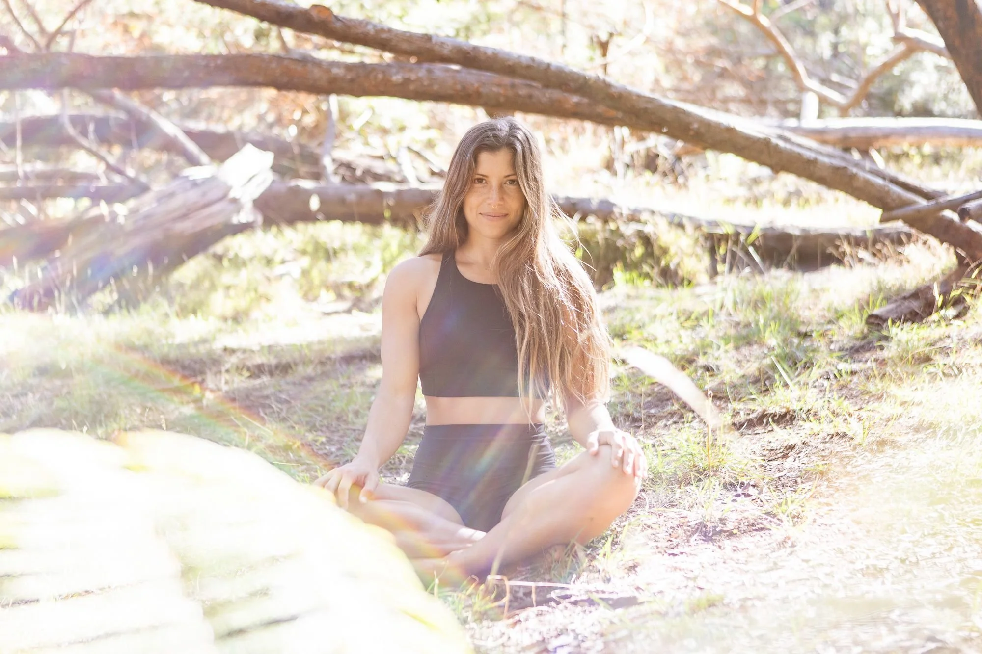 A young woman with long brown hair practicing yoga outdoors in a forest, sitting cross-legged and holding an ankle while smiling at the camera.