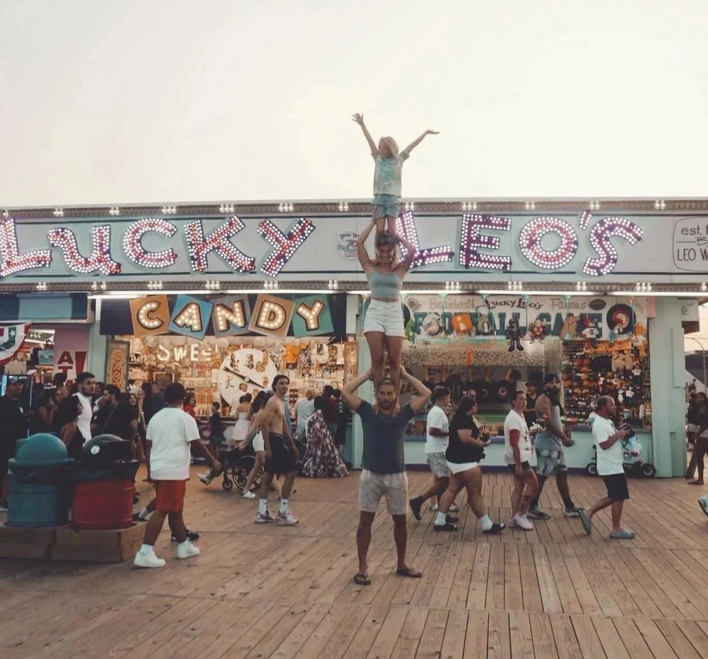 A group of people at a fairground, with a man and child performing a human pyramid in front of an amusement stand. The stand has colorful signs reading 'Lucky Leo's' and 'Candy' with bright lights, and the background is lively with many visitors walking on wooden flooring.