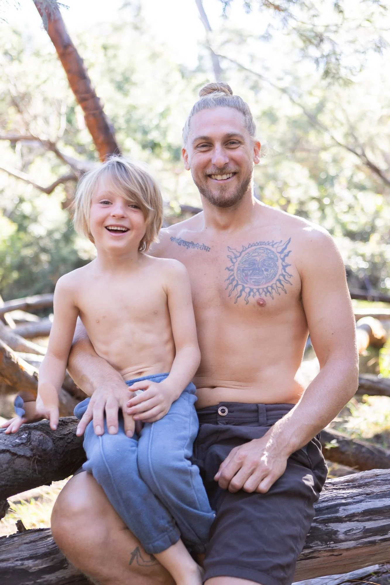 A shirtless man with tattoos smiling and sitting outdoors with a young boy on his lap, both on a fallen log in a forested area with sunlight filtering through the trees.