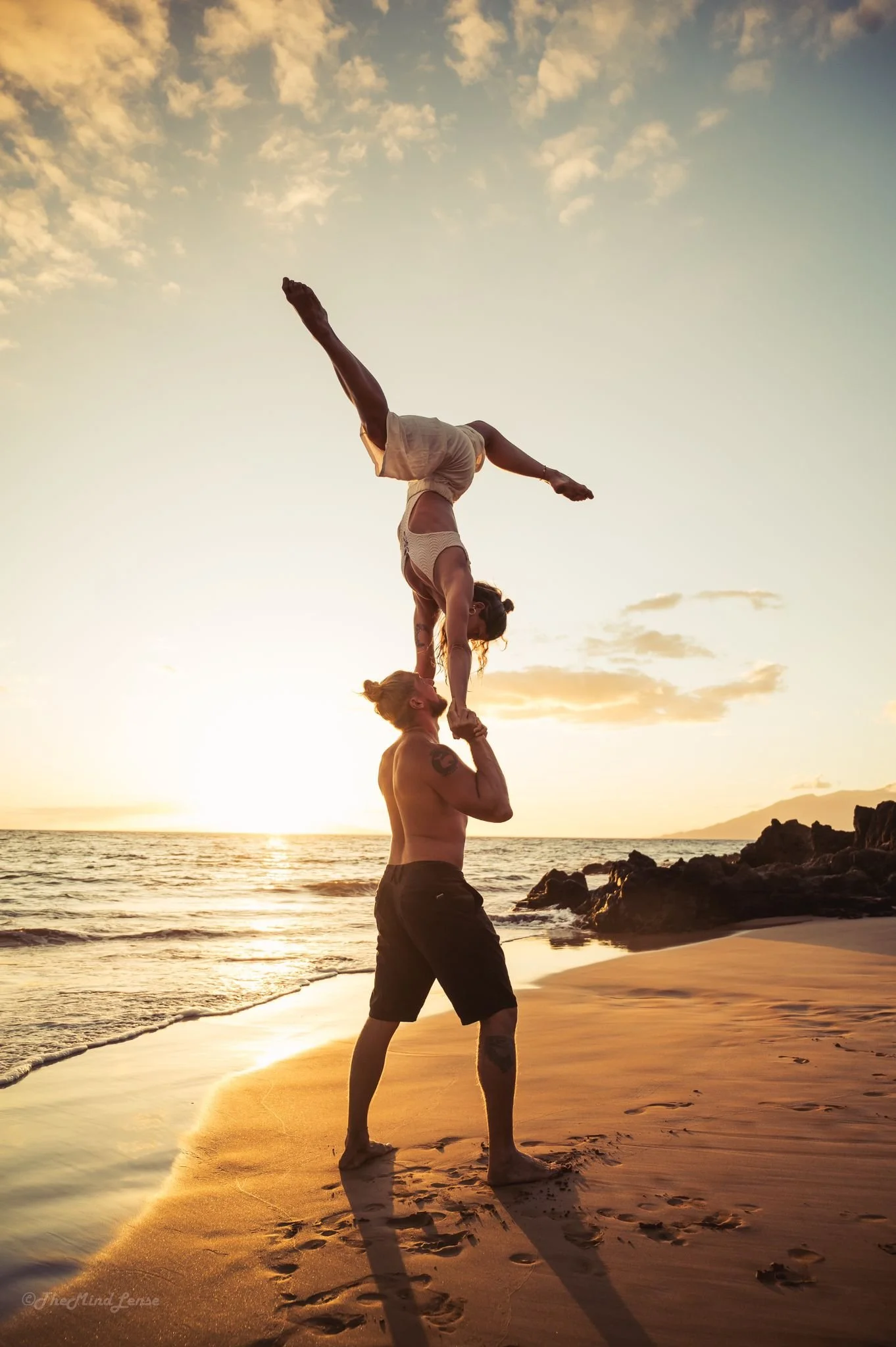 Two people performing a yoga handstand on a beach at sunset. One person with tattooed arms is supporting another person who is upside down, reaching towards the sky with one leg extended and the other bent. The scene features a calm ocean, rocks, and a partly cloudy sky with warm golden light.