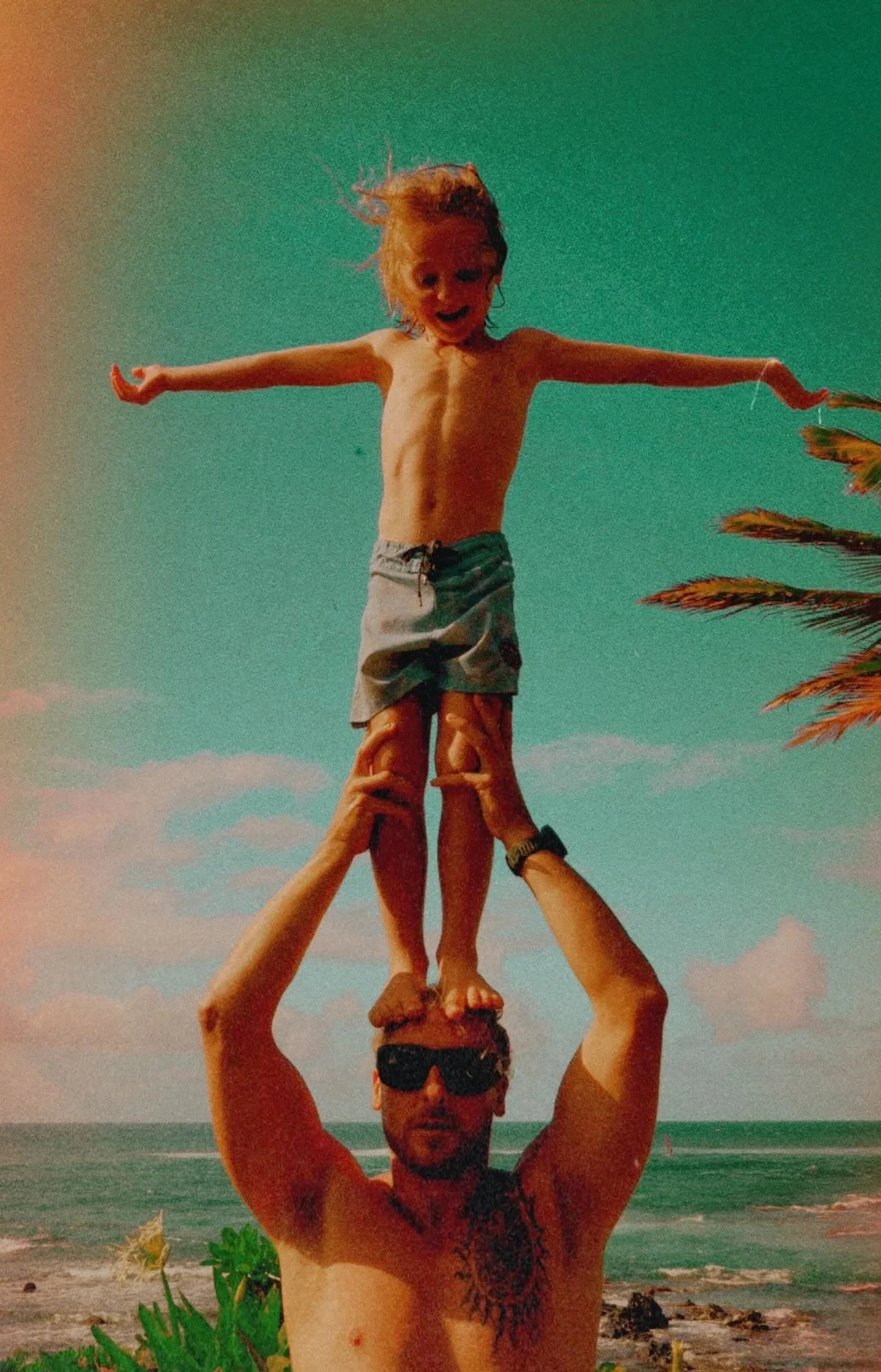 A group of four people on a beach forming a human pyramid with one person standing on top. The top person is a child with their arms outstretched, smiling, and wearing shorts. The sky is partly cloudy, the beach has green plants, palm trees, and the ocean in the background.