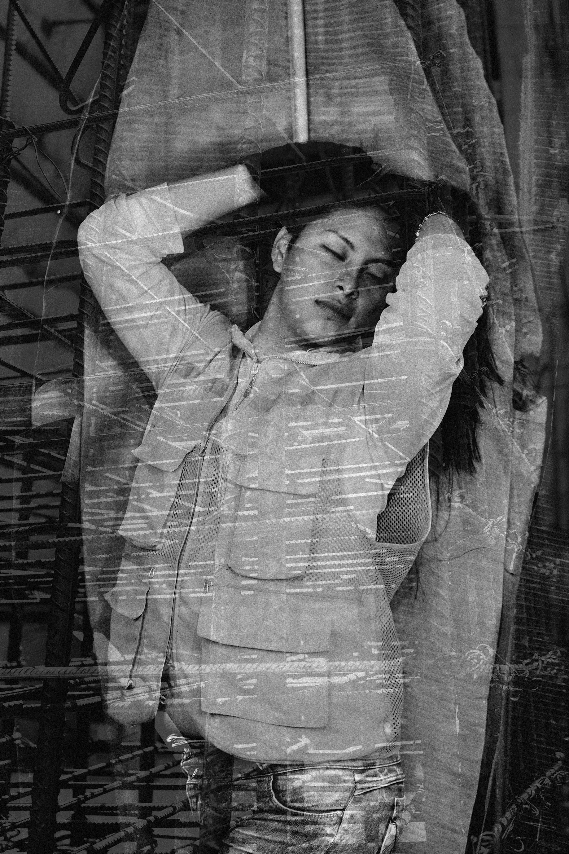 Mujer joven con expresión tranquila, descansando con los brazos detrás de la cabeza, a través de un reflejo en una ventana, en blanco y negro.