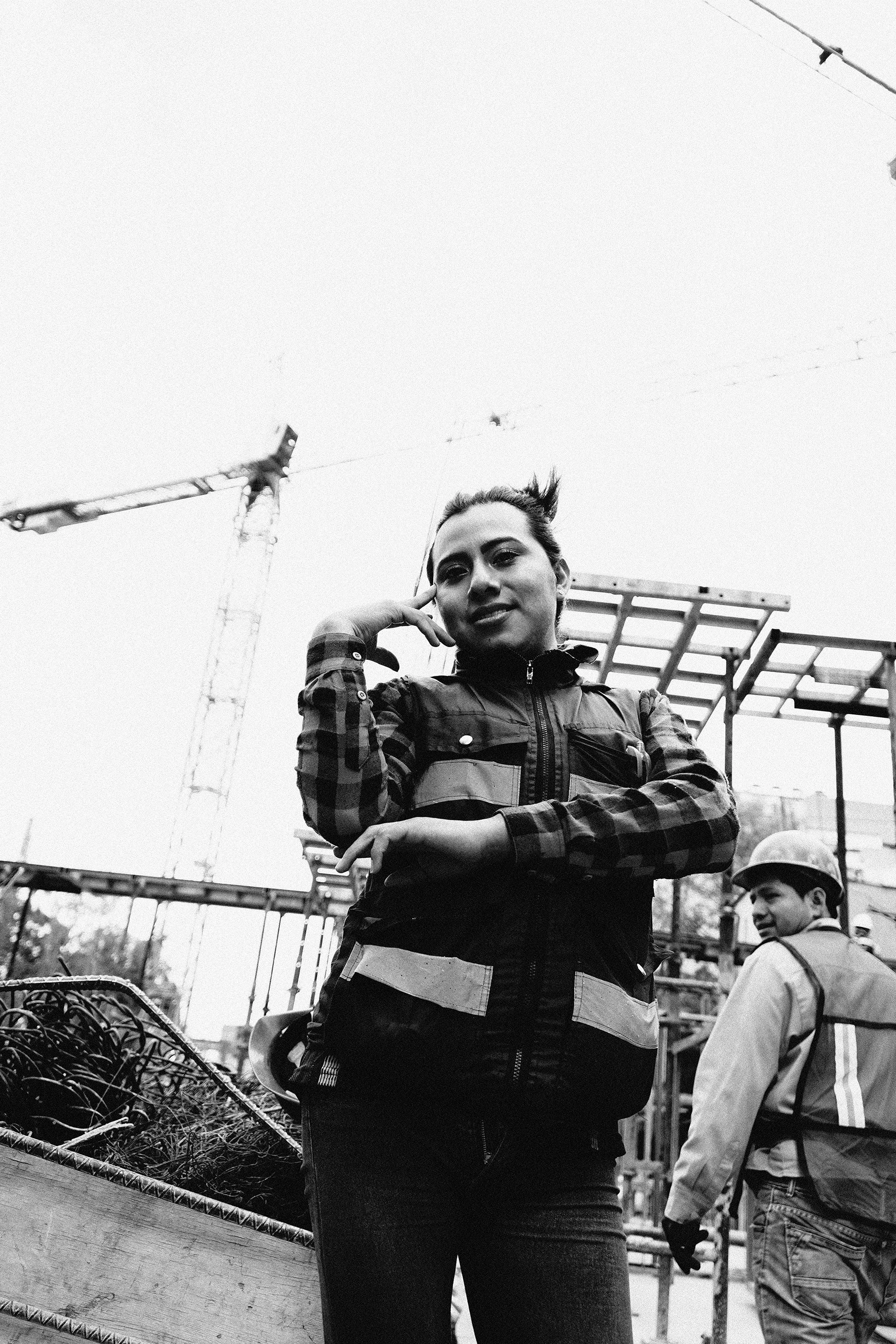 Mujer en sitio de construcción con maquinaria y estructura de metal, otro trabajador con casco en el fondo, en blanco y negro.