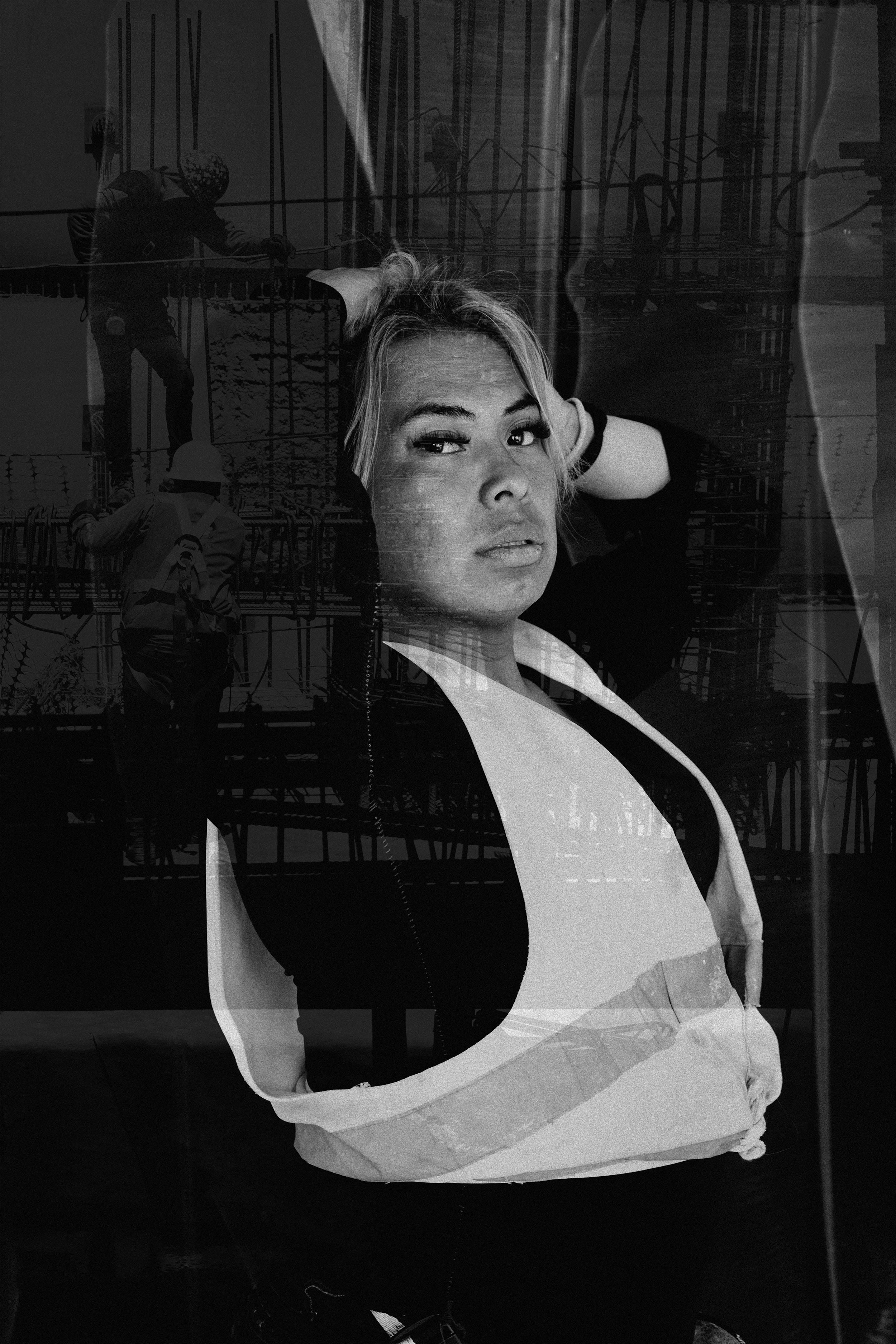 Retrato en blanco y negro de una mujer con expresión pensativa, vista a través de un reflejo que muestra elementos de construcción y trabajadores en el fondo.