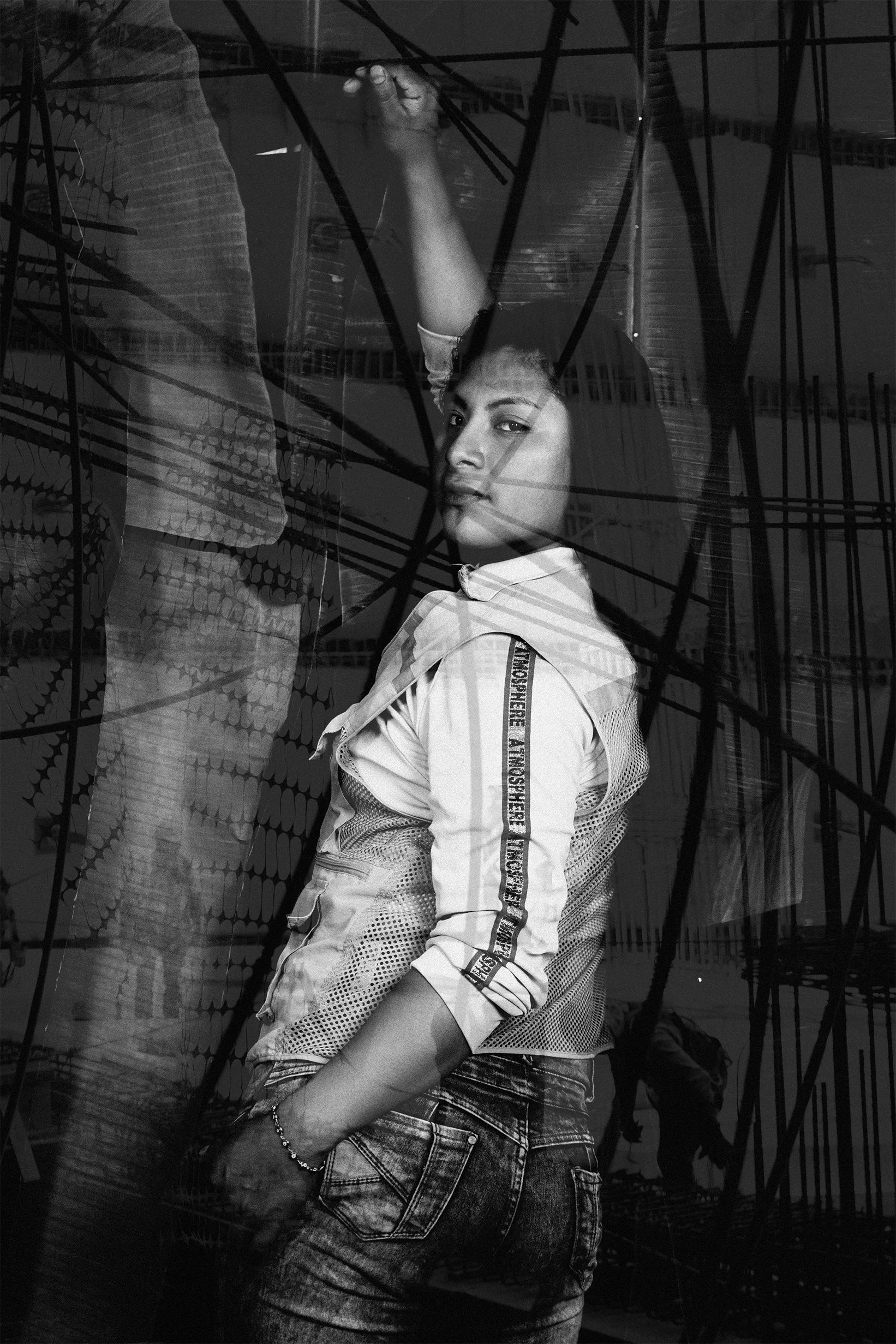 Retrato en blanco y negro de una mujer en una pose artística, con una estructura metálica de fondo y reflejos en el vidrio