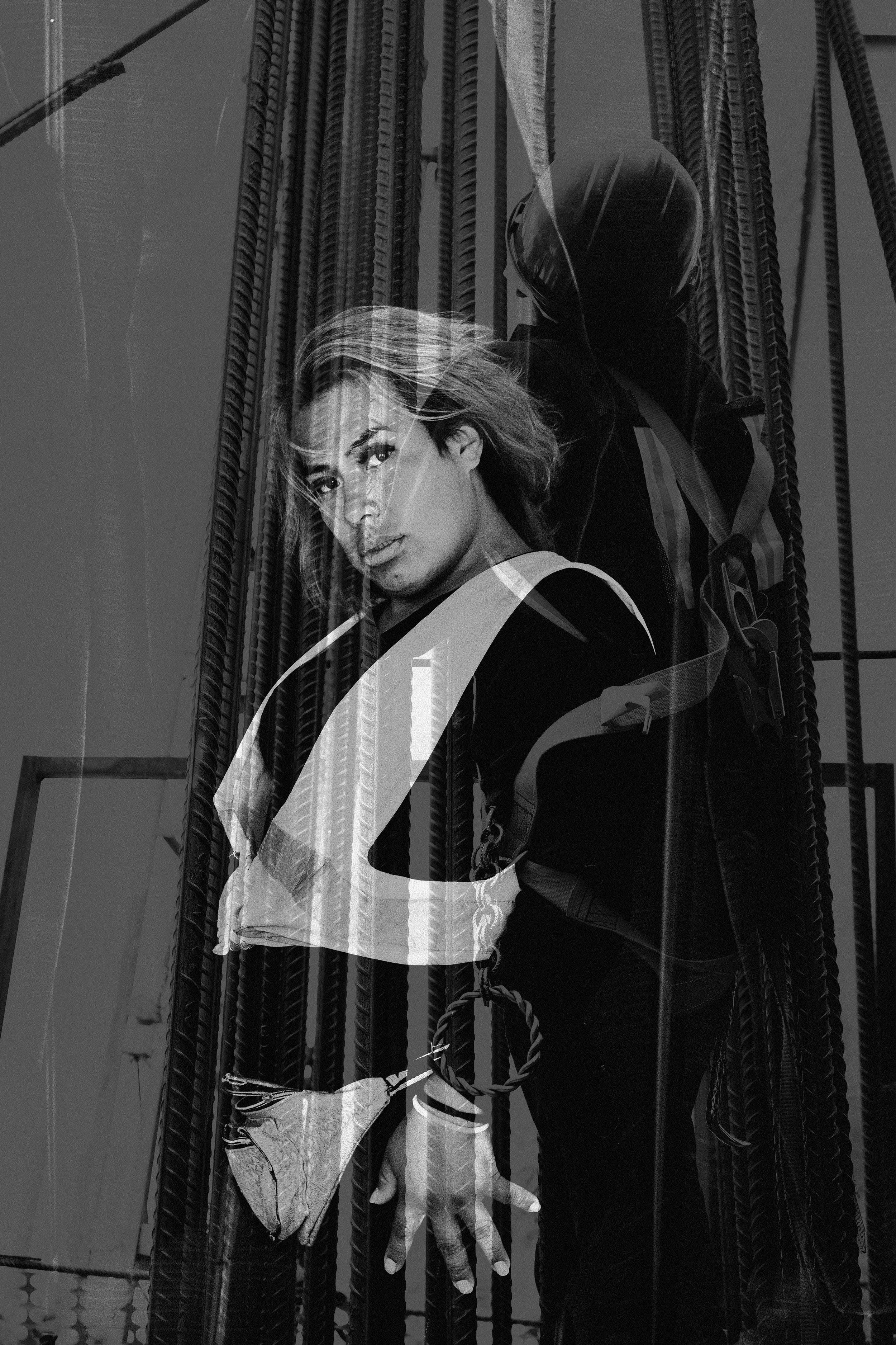 Mujer con cabello rubio y vestido oscuro, viendo hacia la cámara, reflejada en una ventana con estructura metálica.