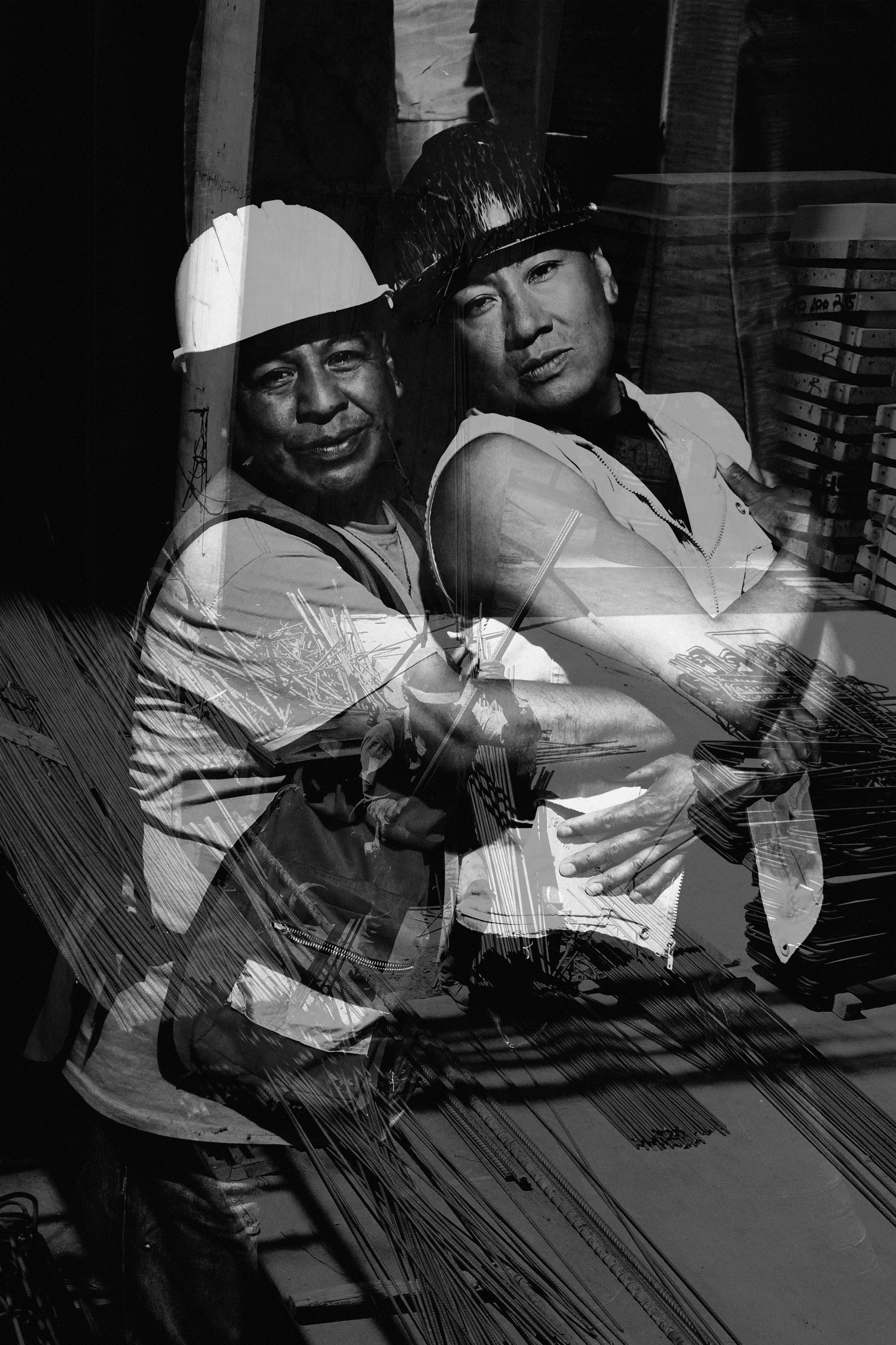 Reflejo en un vidrio de dos hombres con cascos y herramientas, con fondo de pilas de objetos y estructura metálica.