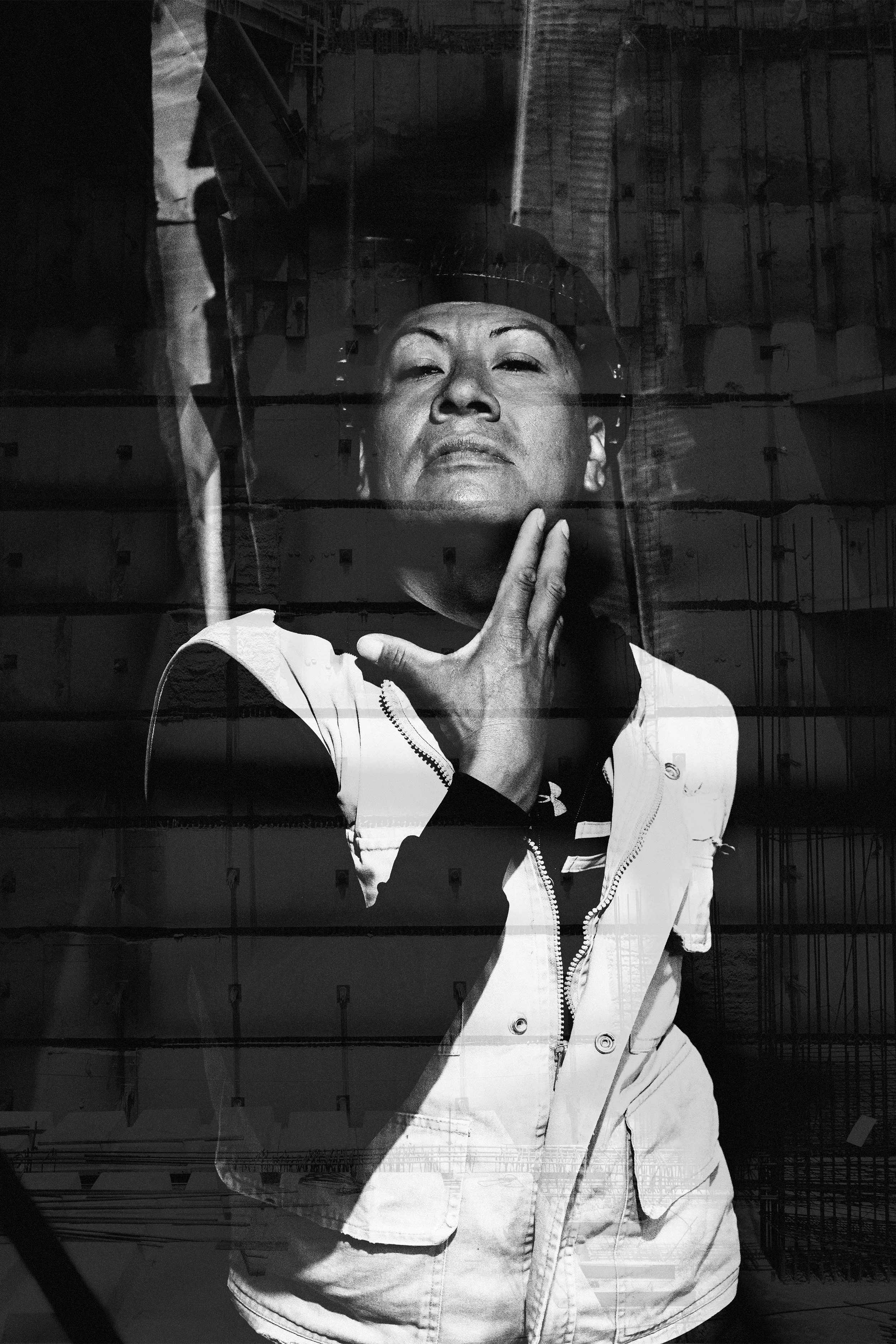 Retrato en blanco y negro de una mujer con expresión confiada y pose pensativa, viendo hacia adelante, con fondo de edificio y estructura metálica.