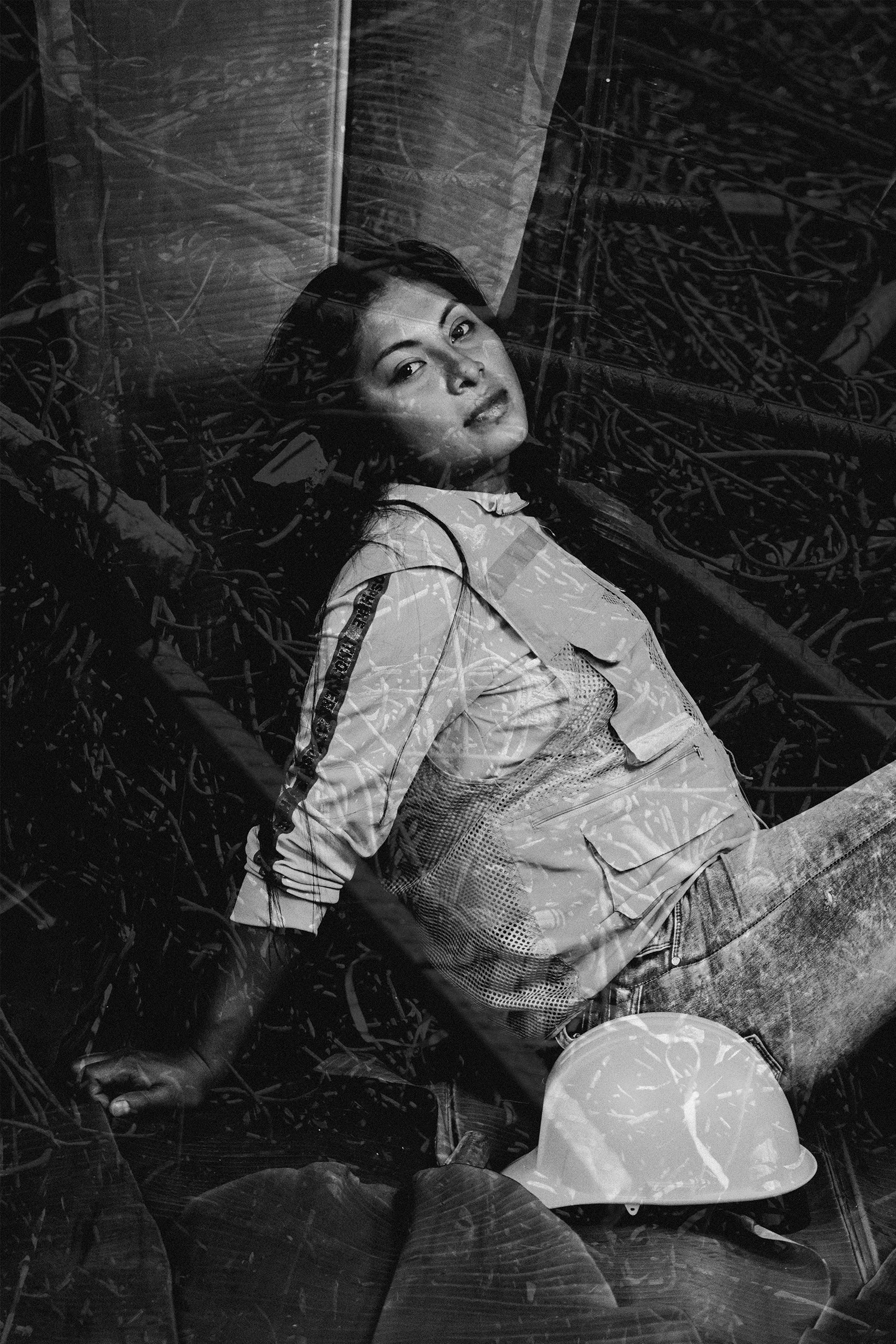 Mujer con uniforme militar recostada sobre maderas y cables, con casco de construcción al frente, vista a través de una ventana o reflejo